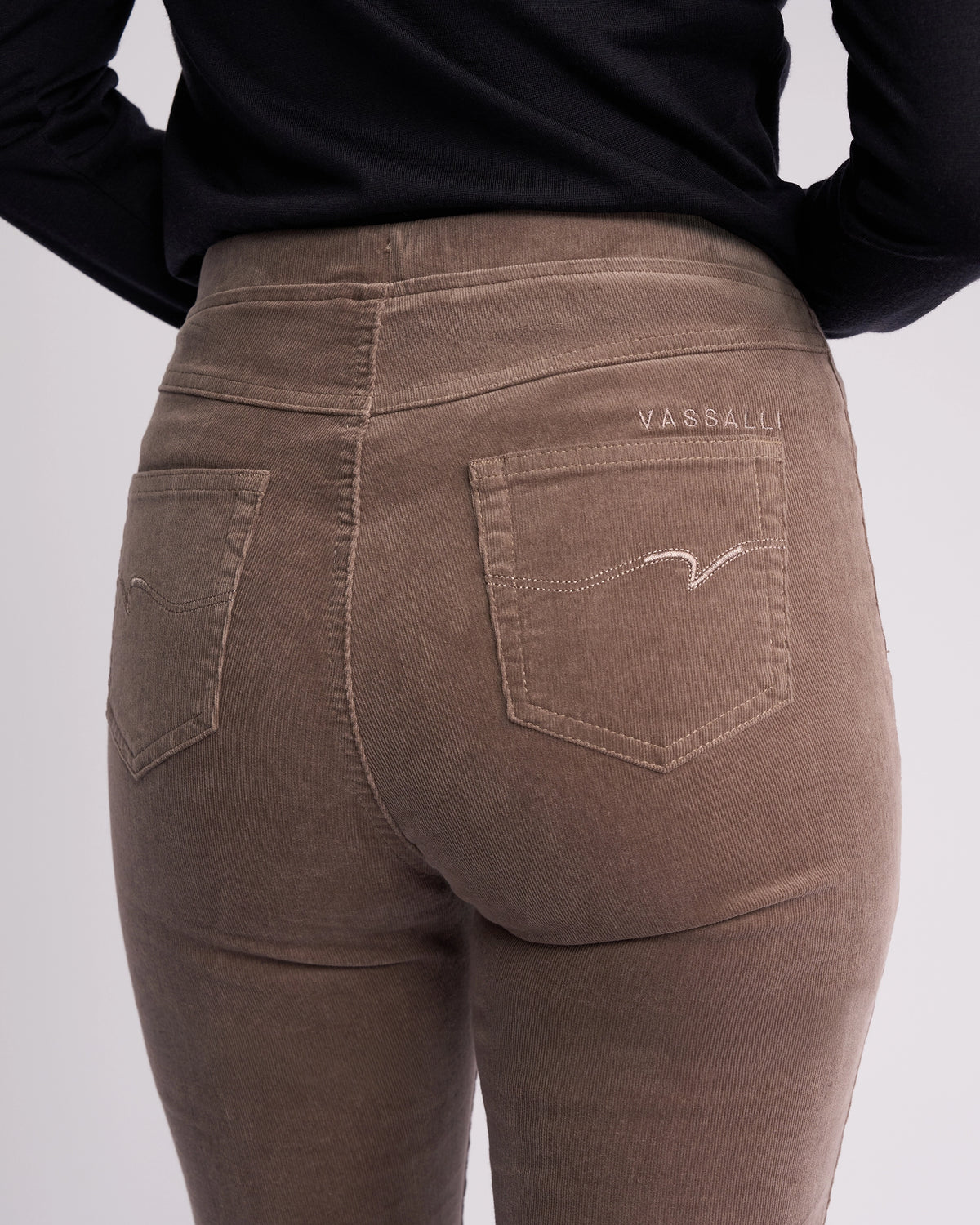 Close-up of a person wearing the Vassalli Bay Cord Pull On in taupe with a black top, highlighting the back pocket featuring Vassalli embroidery above it.