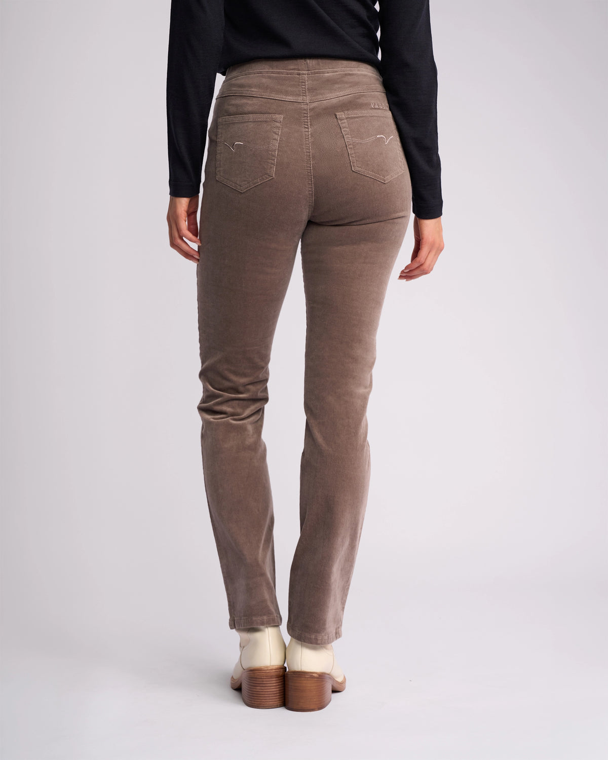A person wearing Vassalli's Bay Cord Pull On pants in taupe, a black long-sleeve top, and cream heeled shoes stands with their back to the camera against a plain light background.