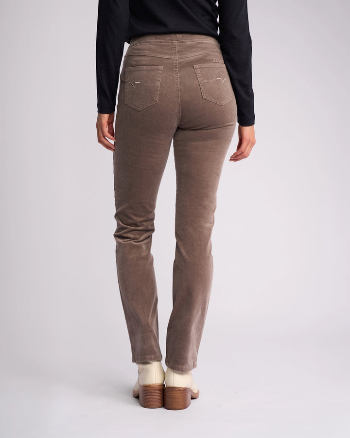 A person wearing Vassalli's Bay Cord Pull On pants in taupe, a black long-sleeve top, and cream heeled shoes stands with their back to the camera against a plain light background.