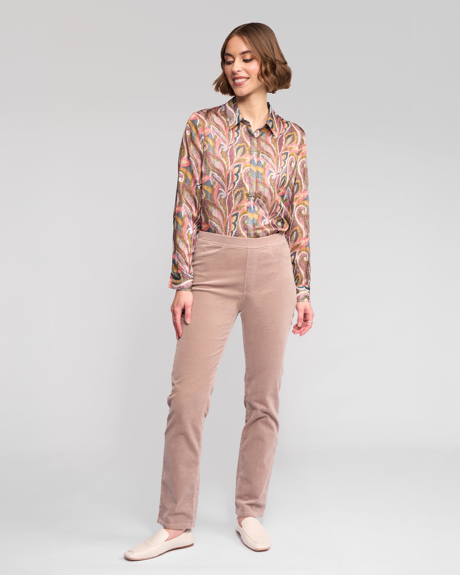 A woman stands smiling, wearing Vassalli Bay Cord Pull On pants in Twig, a patterned long-sleeve shirt, and cream-colored loafers against a plain light gray background.