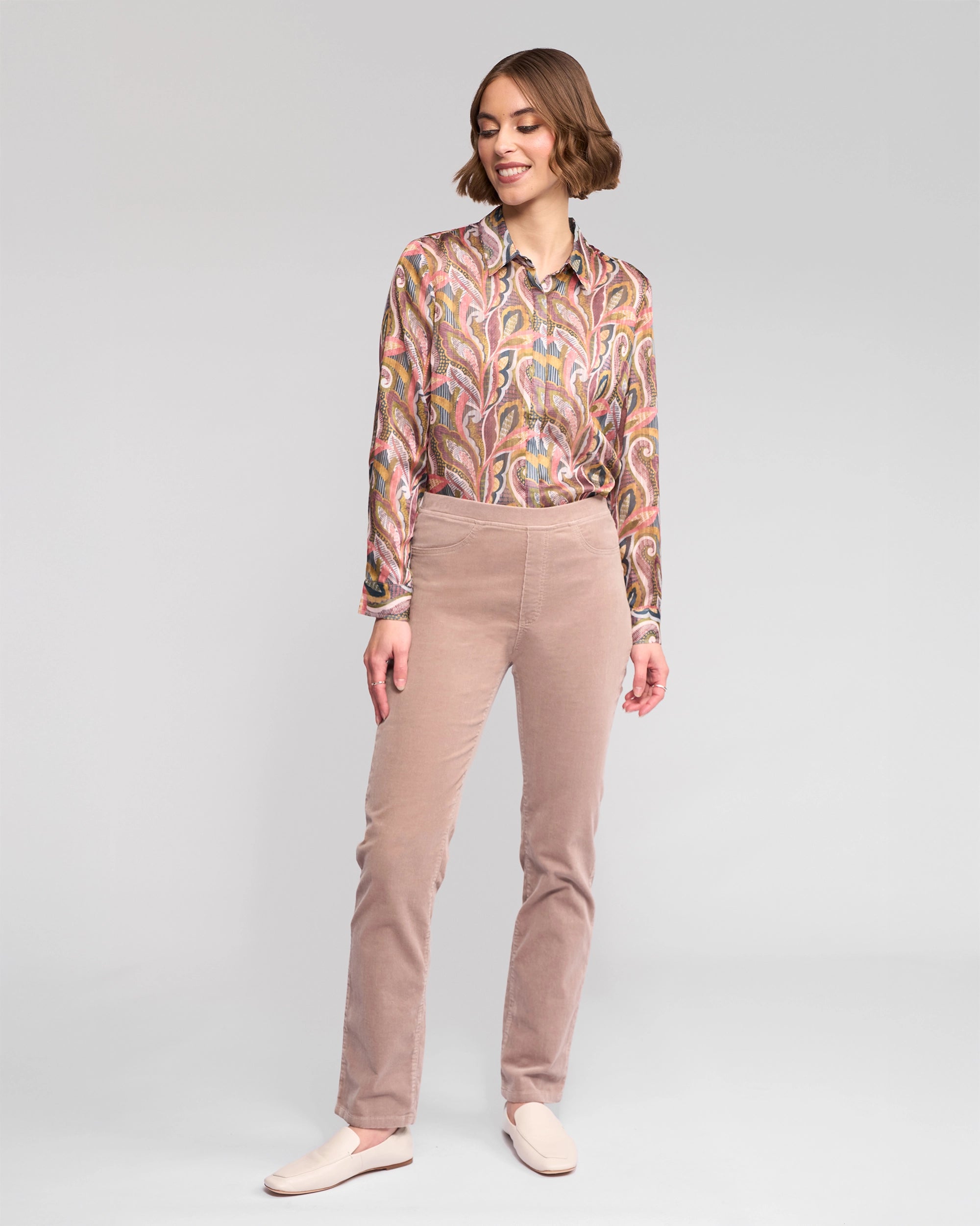 A woman stands against a plain background, smiling in Vassalli's Bay Cord Pull On - Twig pants. She pairs the high-rise slim-leg, light beige trousers with a patterned long-sleeve shirt and matching slip-on shoes, one hand relaxed and the other slightly bent.