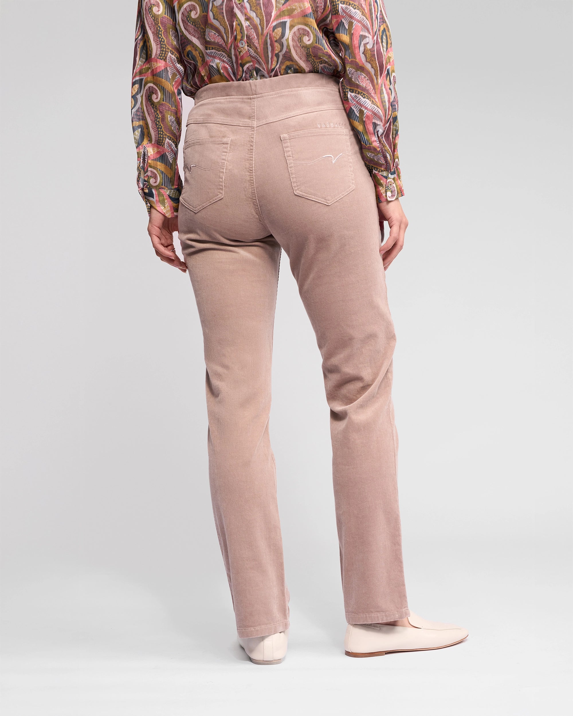 A person wearing Vassalli Bay Cord Pull On - Twig pants stands with their back to the camera on a plain gray background, paired with a colorful paisley shirt and light pink shoes.