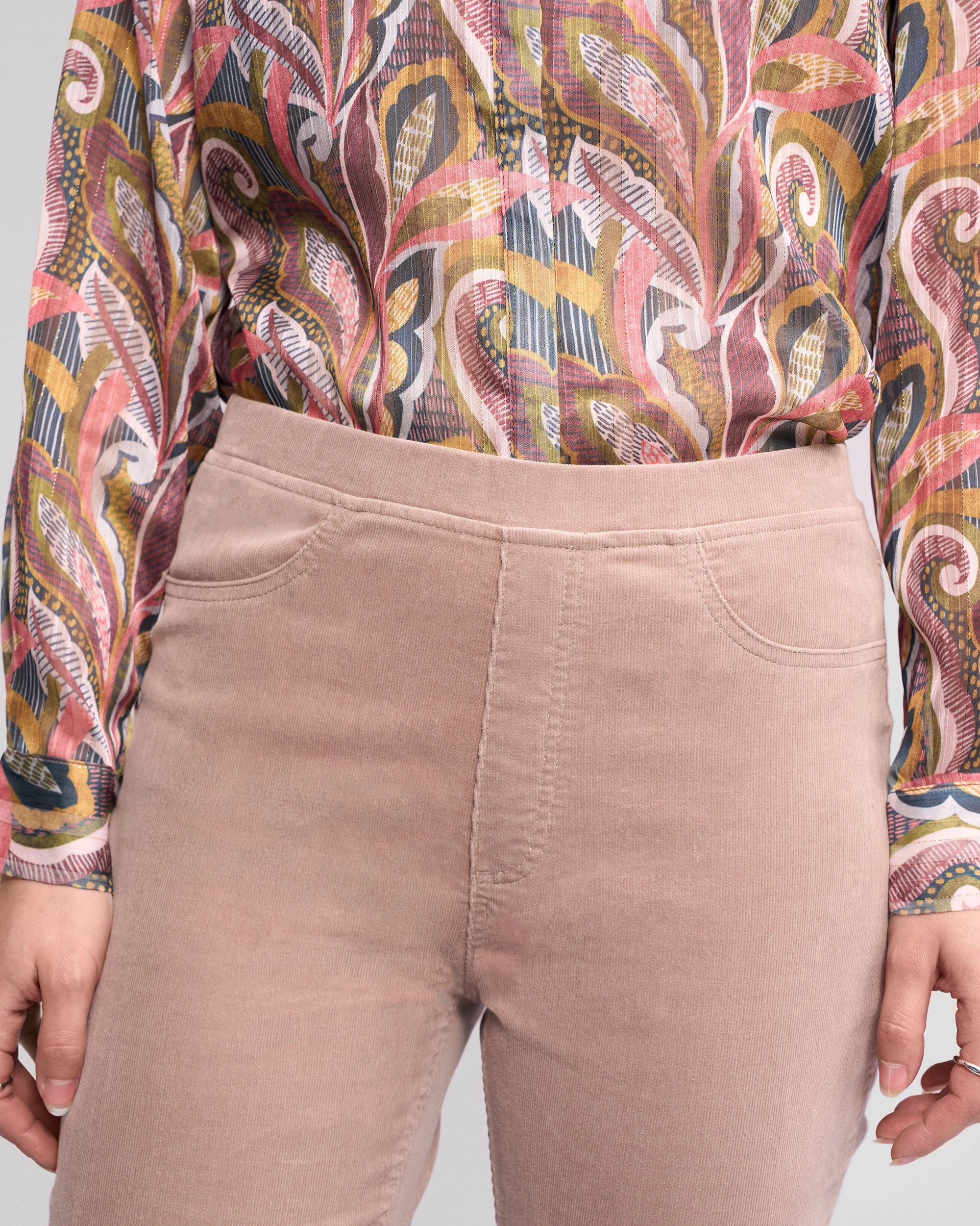 A person wears Vassalli Bay Cord Pull On - Twig high-waisted light pink pants and a long-sleeve shirt with a colorful abstract pattern. The image is cropped from shoulder to mid-thigh.