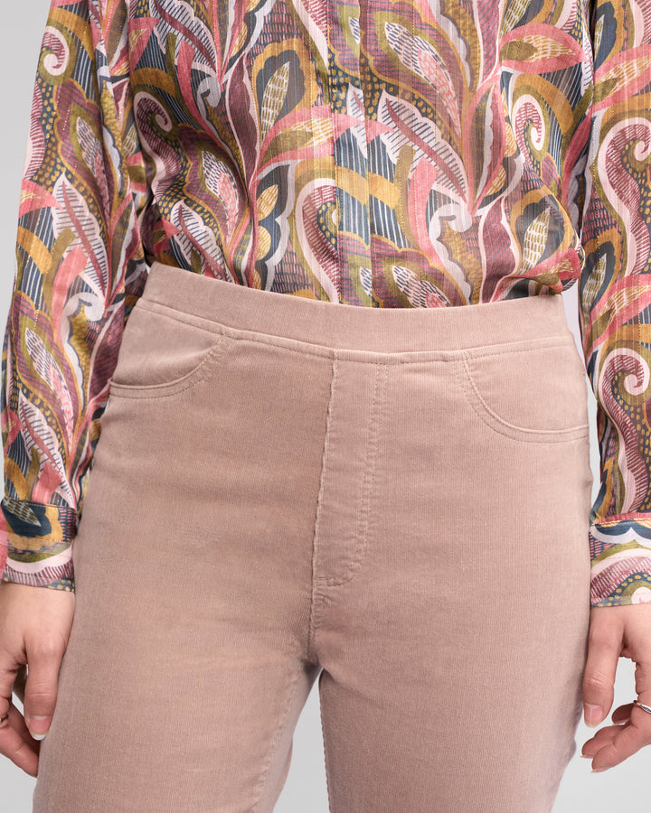 A person wears Vassalli Bay Cord Pull On - Twig high-waisted light pink pants and a long-sleeve shirt with a colorful abstract pattern. The image is cropped from shoulder to mid-thigh.