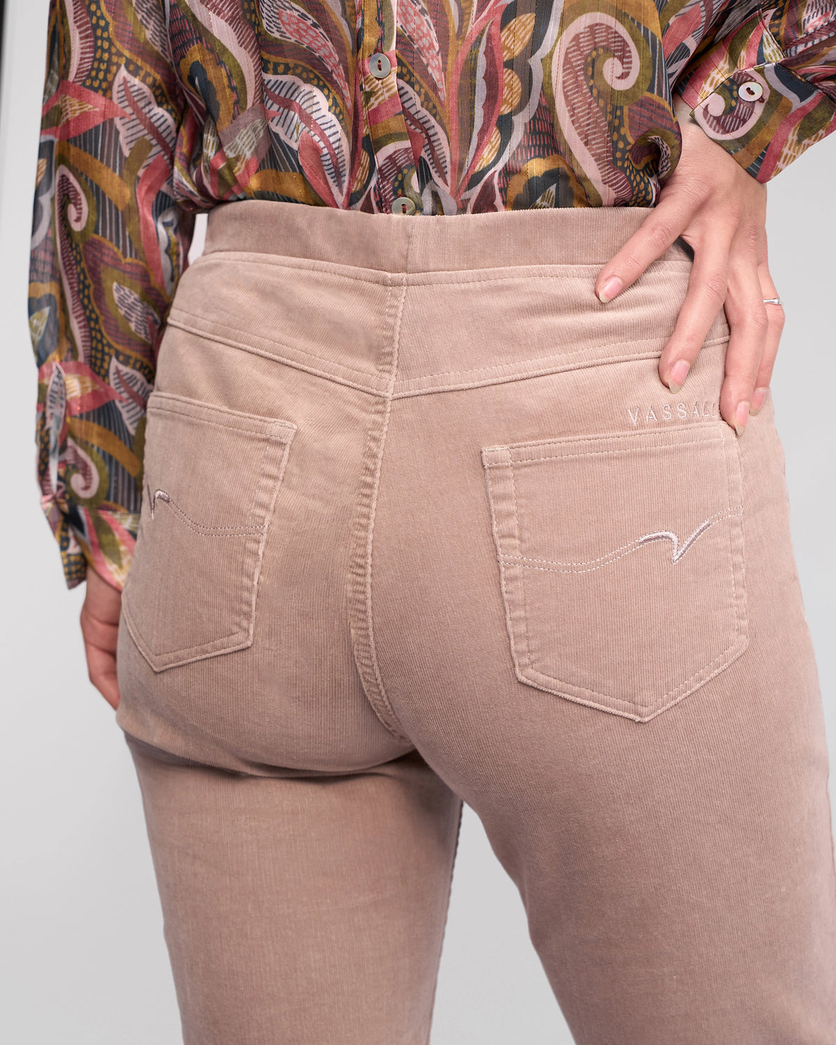 A person models the Vassalli Bay Cord Pull On - Twig pants, featuring beige stretch pinwale cord, slim legs, back pockets with wavy stitching, paired with a colorful patterned shirt. The photo highlights the lower back and hips.