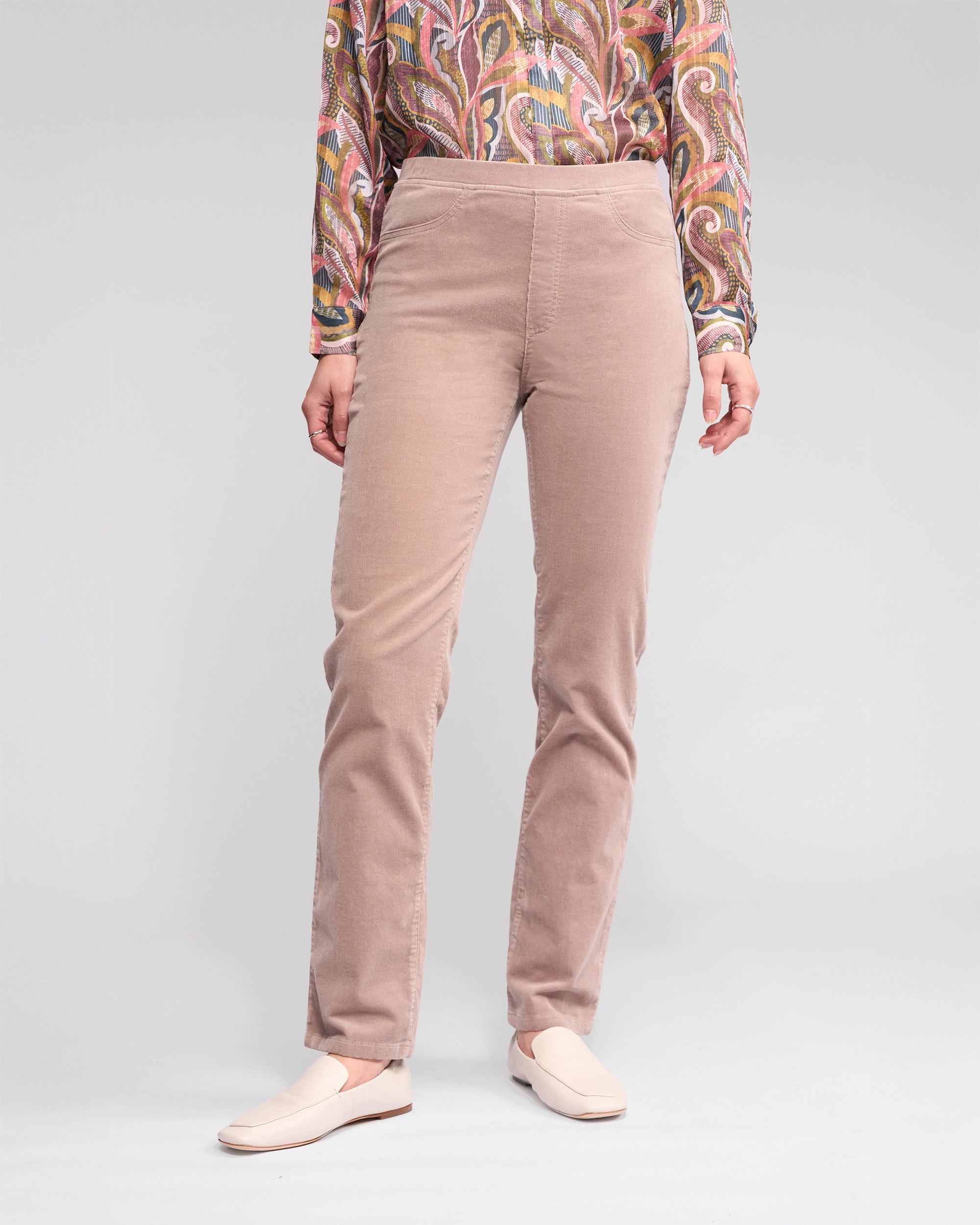 A person wears Vassalli Bay Cord Pull On - Twig pants, a colorful wavy long-sleeve shirt, and cream slip-on shoes, standing against a plain light background. Only the lower half of the body is visible.