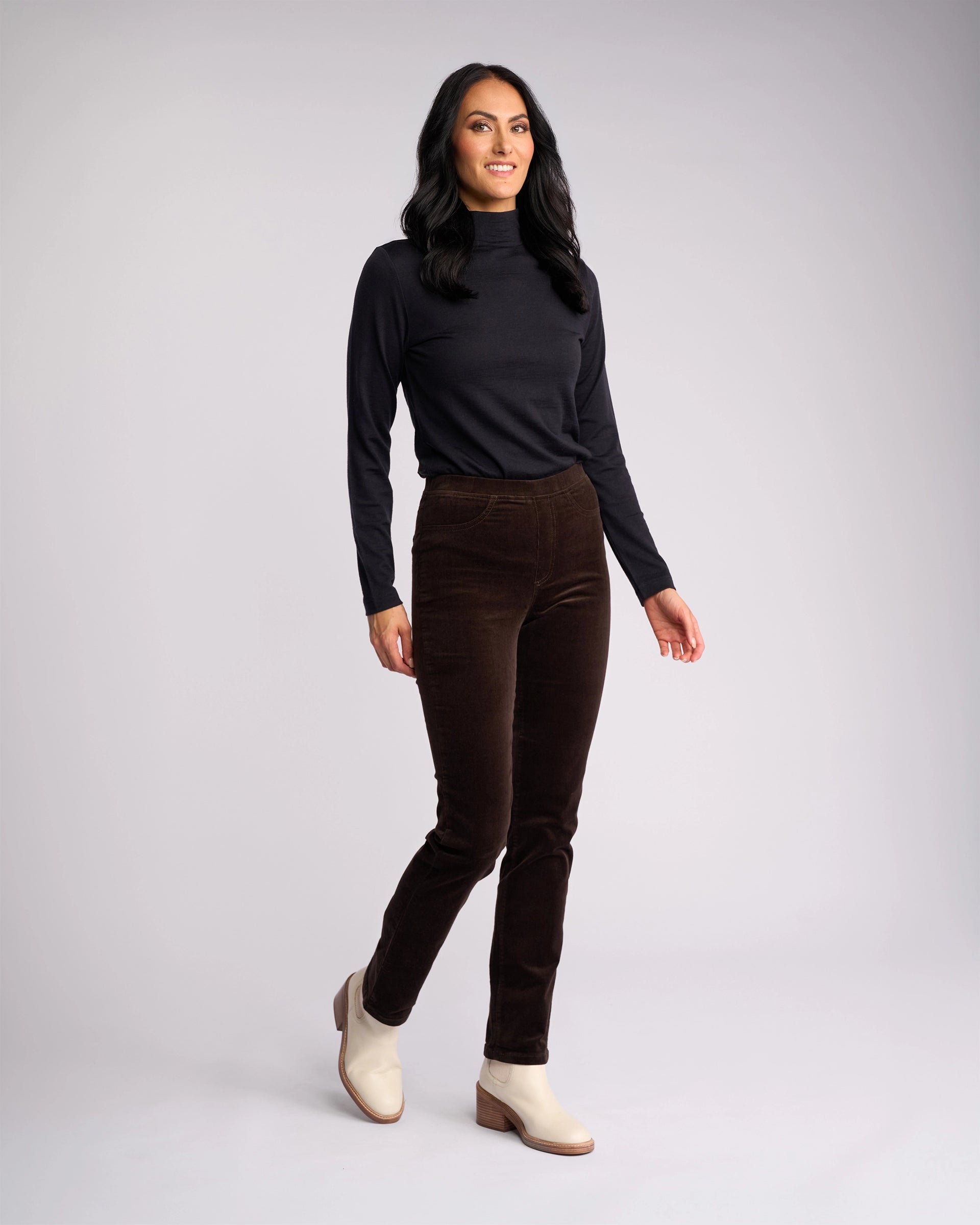 A woman with long dark hair wears a black long-sleeve top, light ankle boots, and Vassalli Bay Cord Pull On pants in Walnut. She stands smiling against a plain light gray background.
