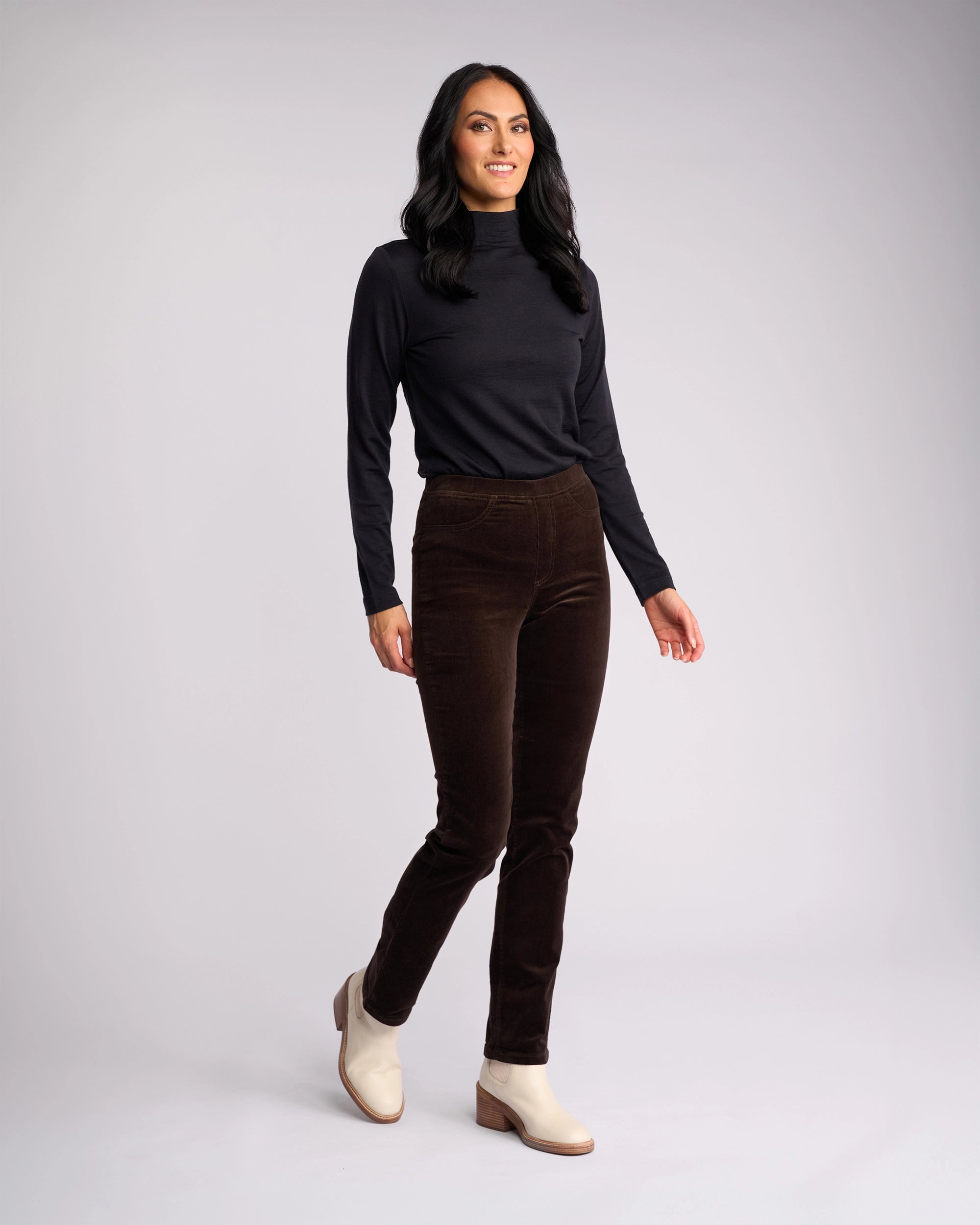 A woman with long dark hair wears a black long-sleeve top, light ankle boots, and Vassalli Bay Cord Pull On pants in Walnut. She stands smiling against a plain light gray background.