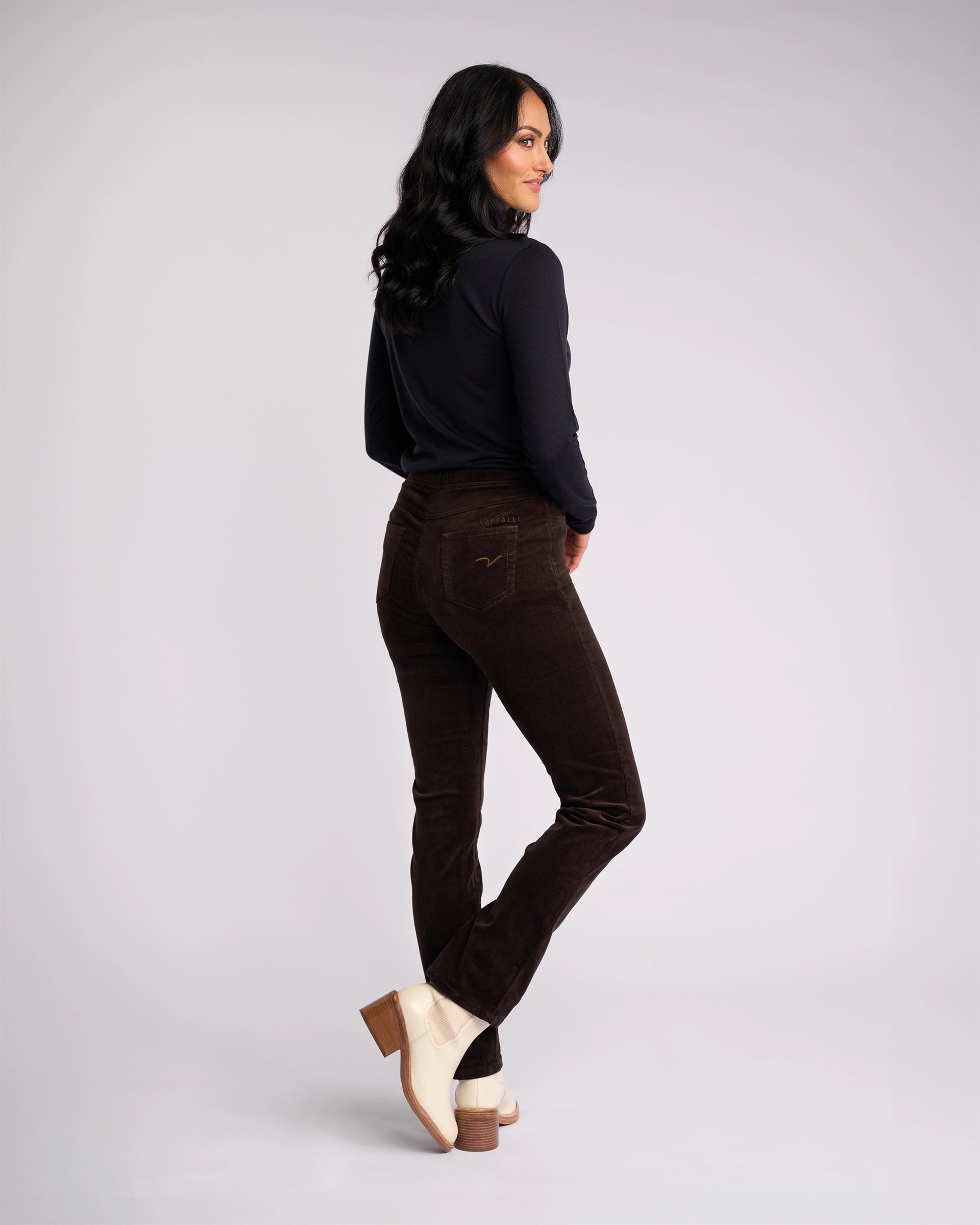 A woman with long dark hair wears a black long-sleeve top and Vassalli Bay Cord Pull On - Walnut pants, paired with beige heeled boots. She stands slightly turned on a white background, smiling.
