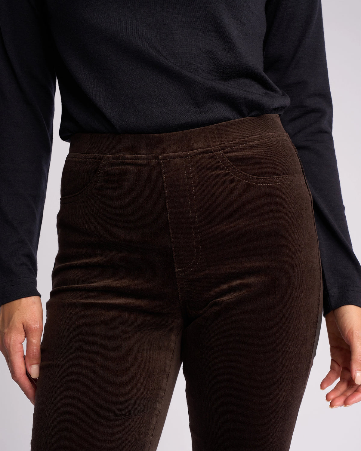 A person wearing a long-sleeved black top and Vassalli Bay Cord Pull On - Walnut slim-leg pants, shown from the shoulders to the upper thighs.