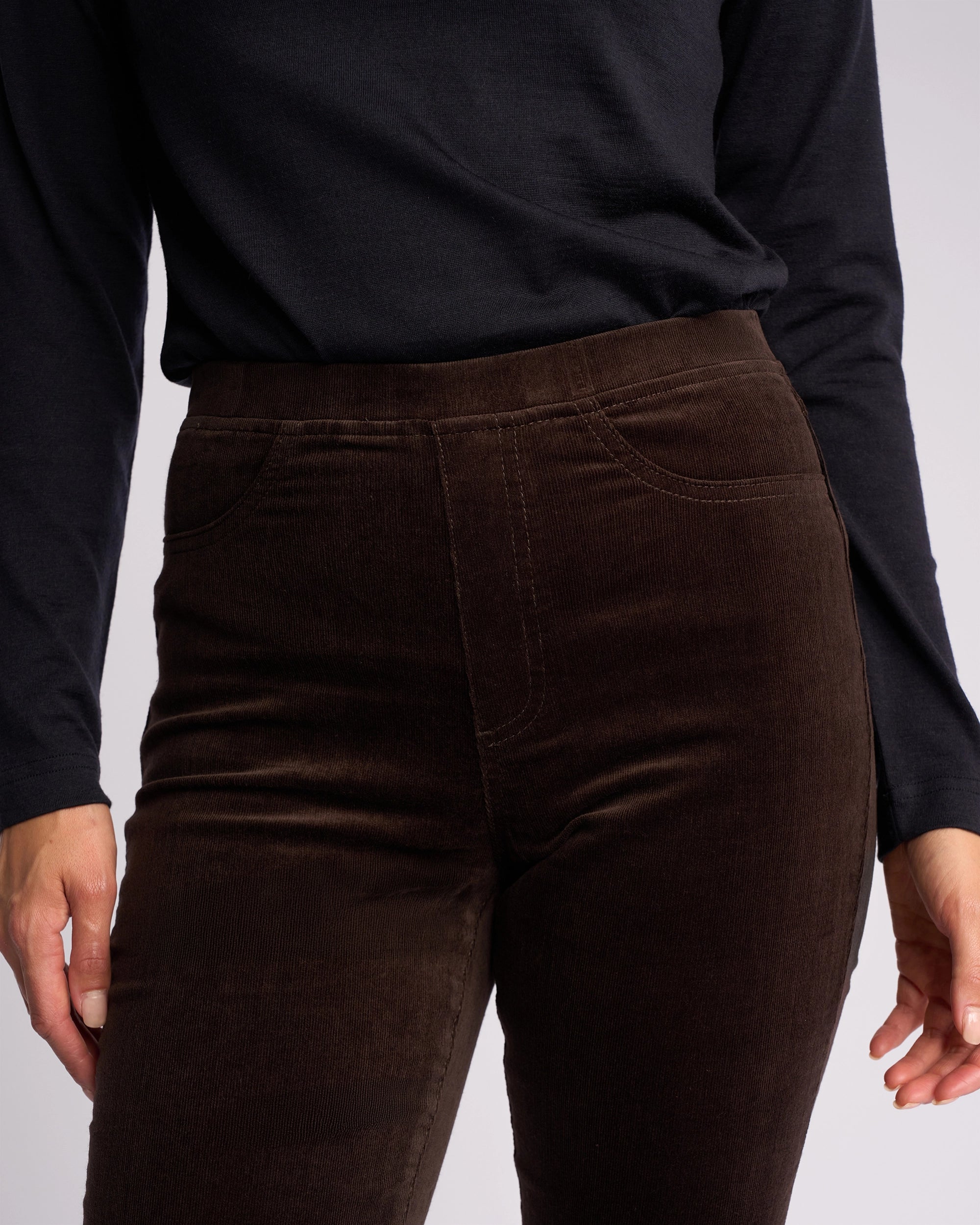 A person wearing a long-sleeved black top and Vassalli Bay Cord Pull On - Walnut slim-leg pants, shown from the shoulders to the upper thighs.