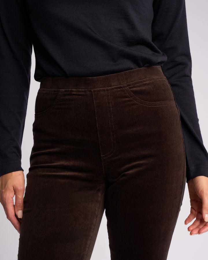 A person wearing a long-sleeved black top and Vassalli Bay Cord Pull On - Walnut slim-leg pants, shown from the shoulders to the upper thighs.