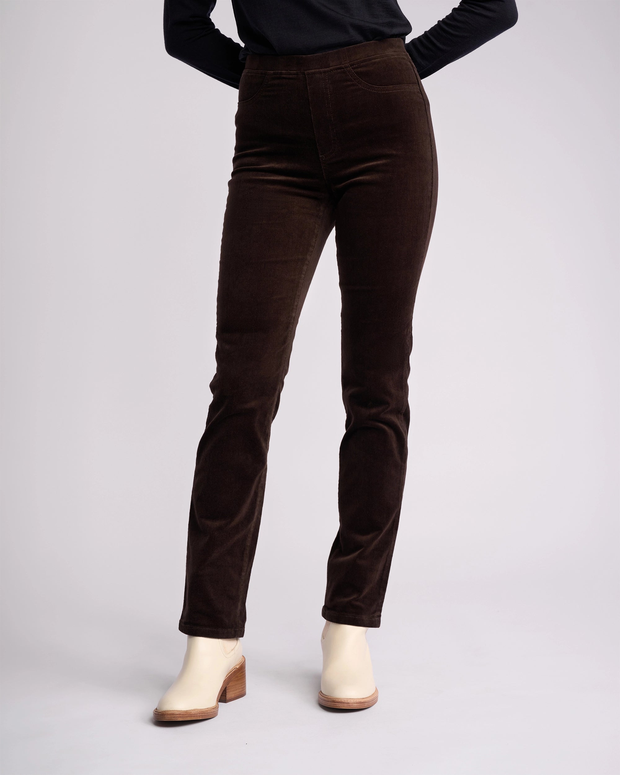 A person wears Vassalli’s Bay Cord Pull On pants in Walnut with cream heeled ankle boots, standing against a plain light background. Only their lower body is visible.