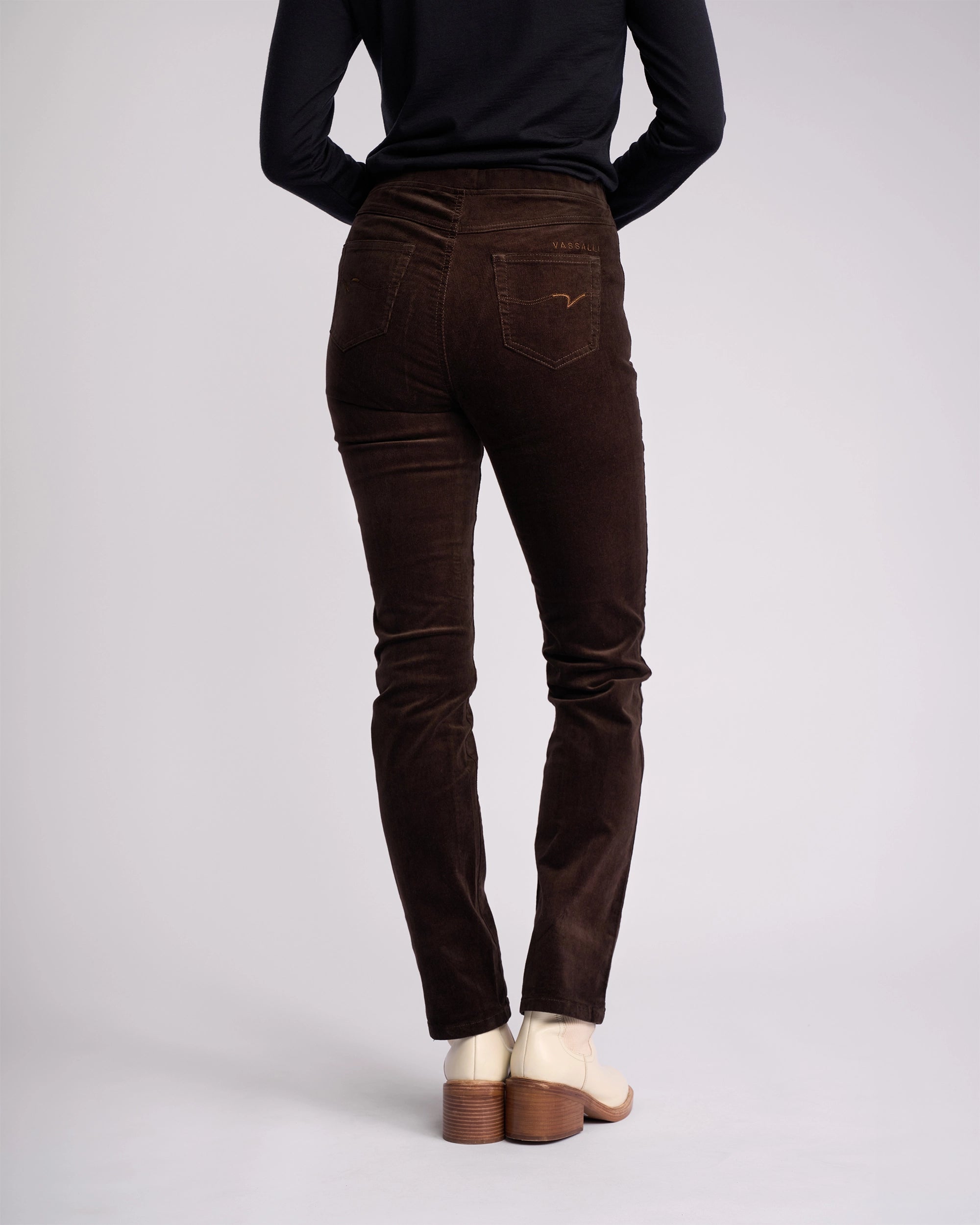 A person is seen from behind wearing a black long-sleeve top, Vassalli Bay Cord Pull On pants in Walnut, and cream heeled shoes, standing against a plain light background.