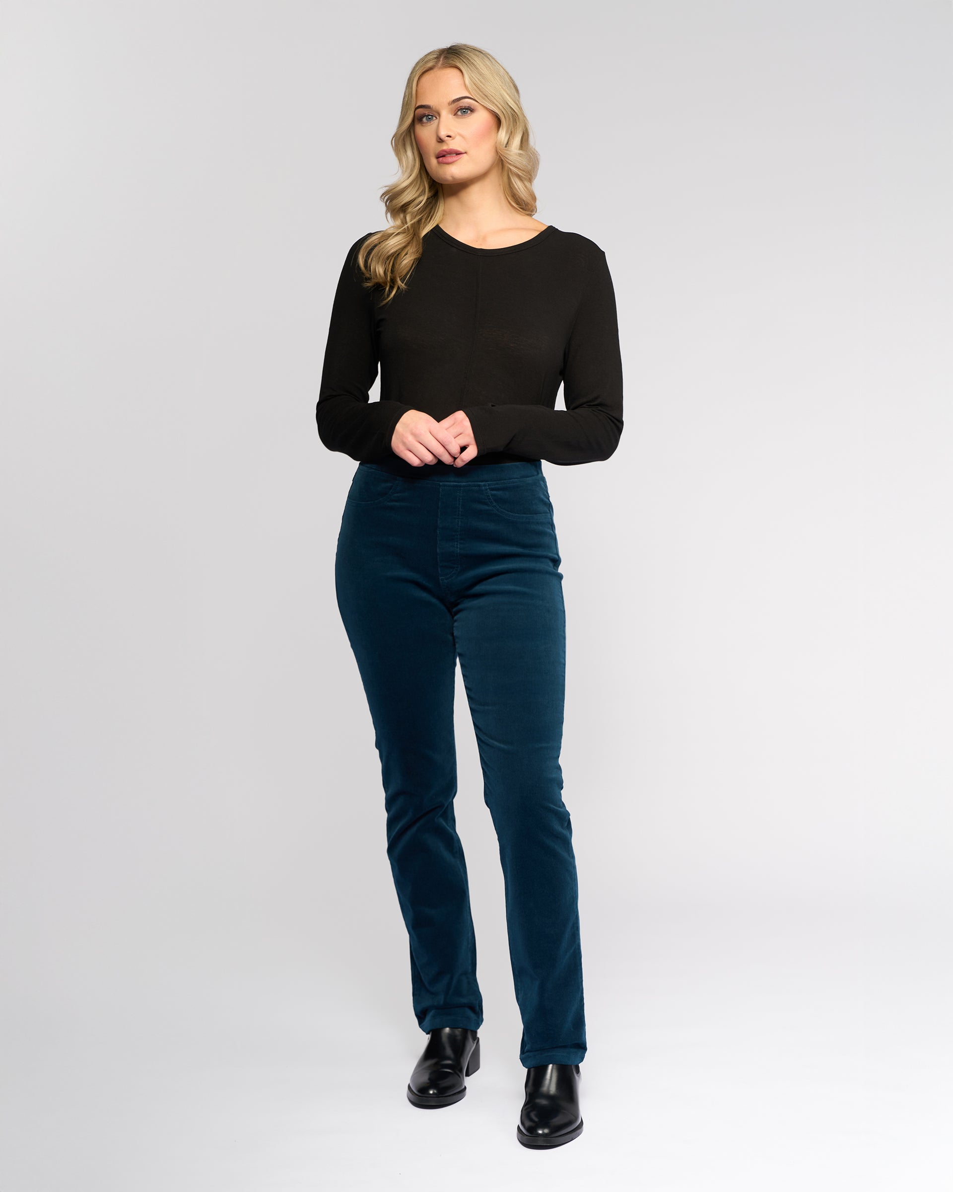 A woman with long blonde hair wears a black long-sleeve top, Vassalli Bay Cord Pull On - Petrol pants, and black shoes, standing against a plain white background.