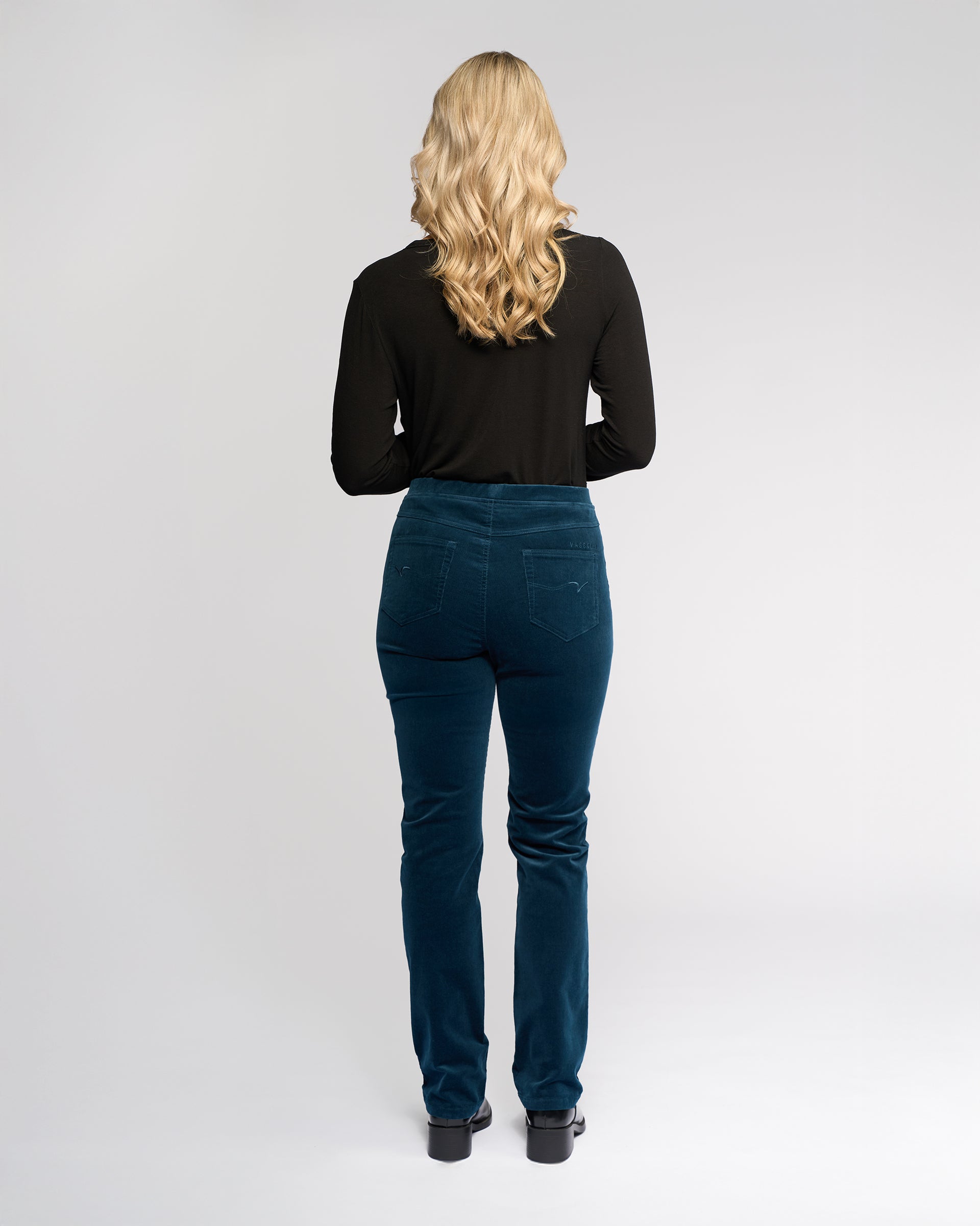 A woman with long blonde hair faces away, wearing the Vassalli Bay Cord Pull On in Petrol—a black long-sleeve top and slim stretch pinwale cord pant. The background is plain and light.