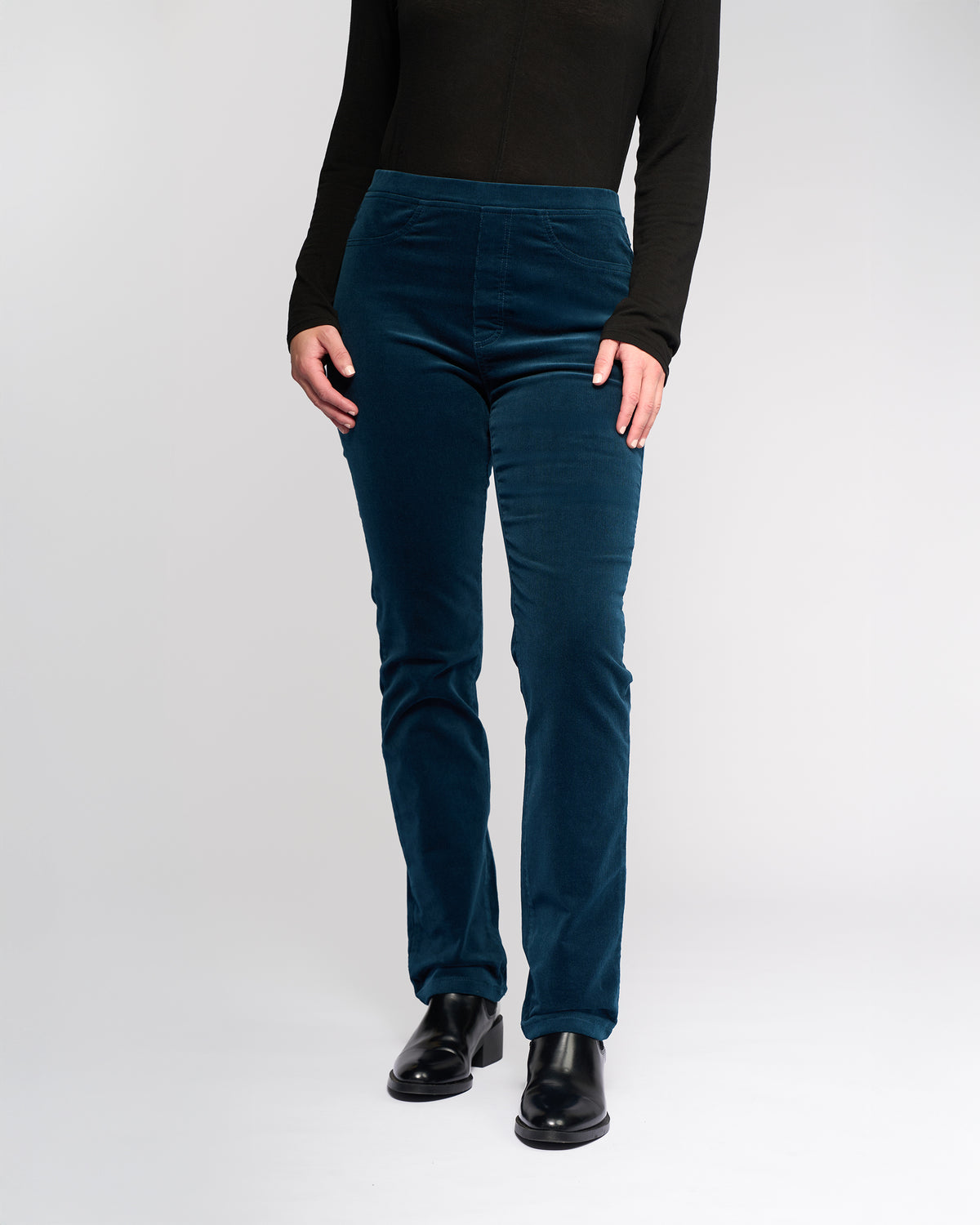 A person wearing a long-sleeve black top, Vassalli Bay Cord Pull On pants in petrol, and black shoes stands against a plain white background. Their head is not visible in the image.