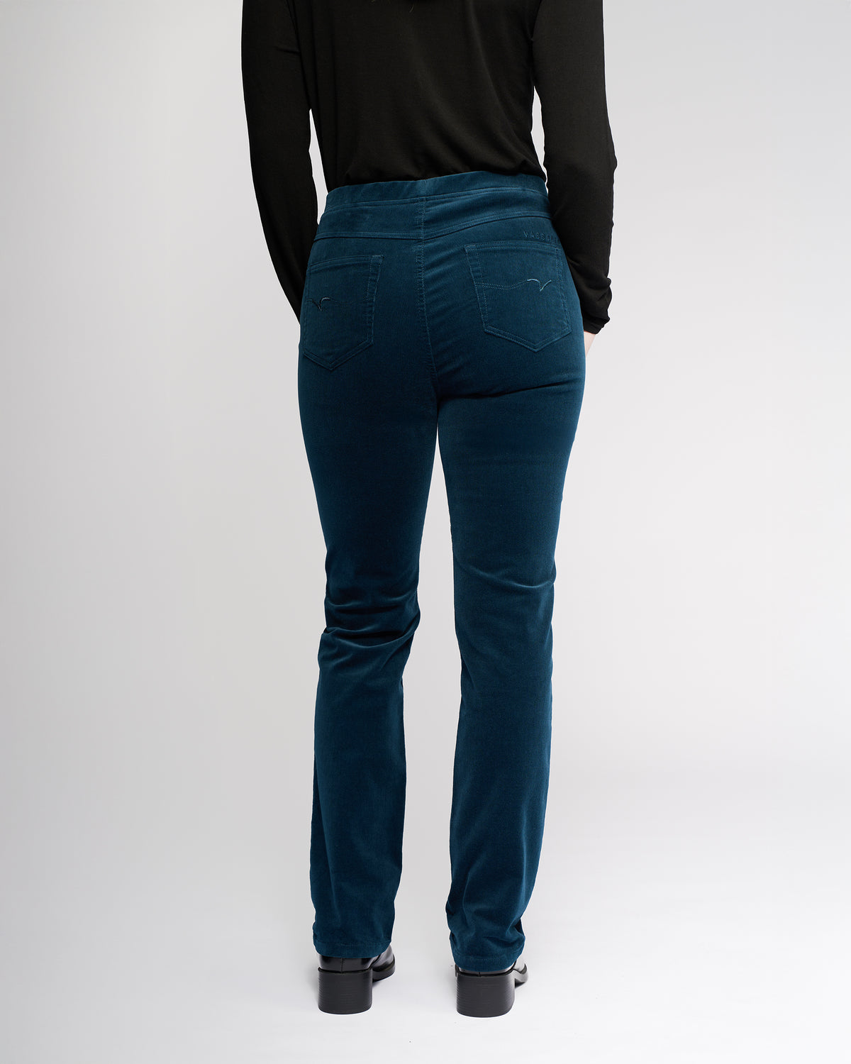 A person wearing a black long-sleeve top and Vassalli Bay Cord Pull On - Petrol pants stands facing away from the camera against a plain light background.