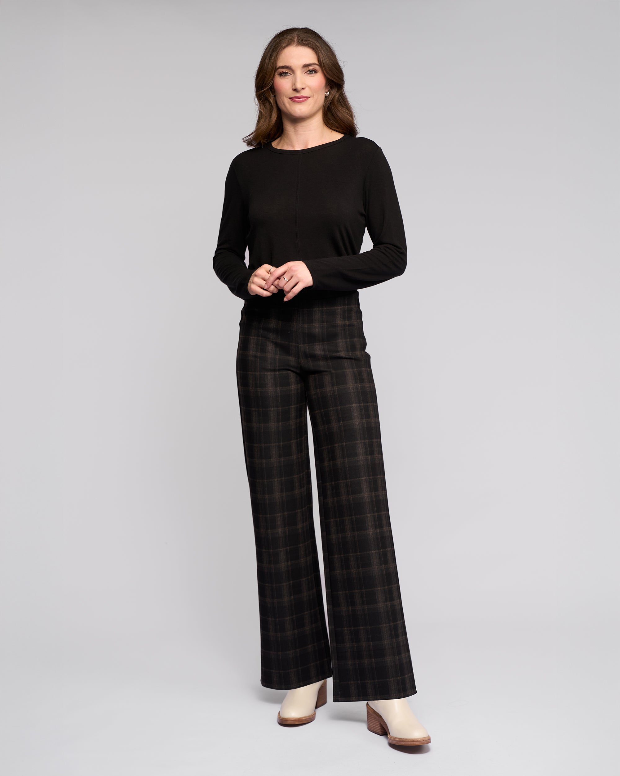 A woman smiles with her hands clasped in front of her, wearing a Vassalli Loft Pull On - Checkmate wide-leg pant, a long-sleeve black top, and cream heeled boots against a plain background.