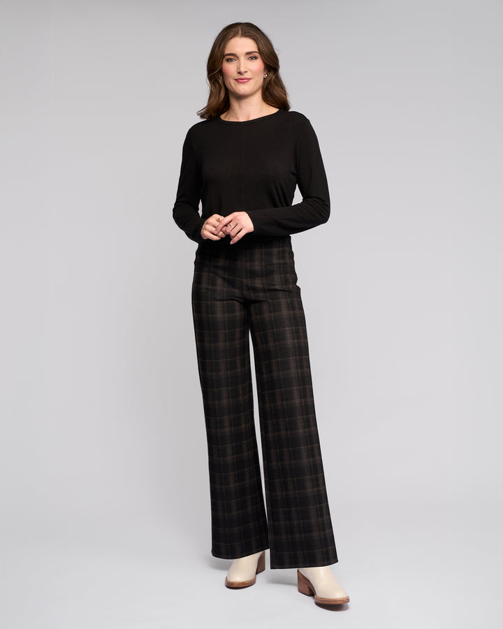 A woman smiles with her hands clasped in front of her, wearing a Vassalli Loft Pull On - Checkmate wide-leg pant, a long-sleeve black top, and cream heeled boots against a plain background.