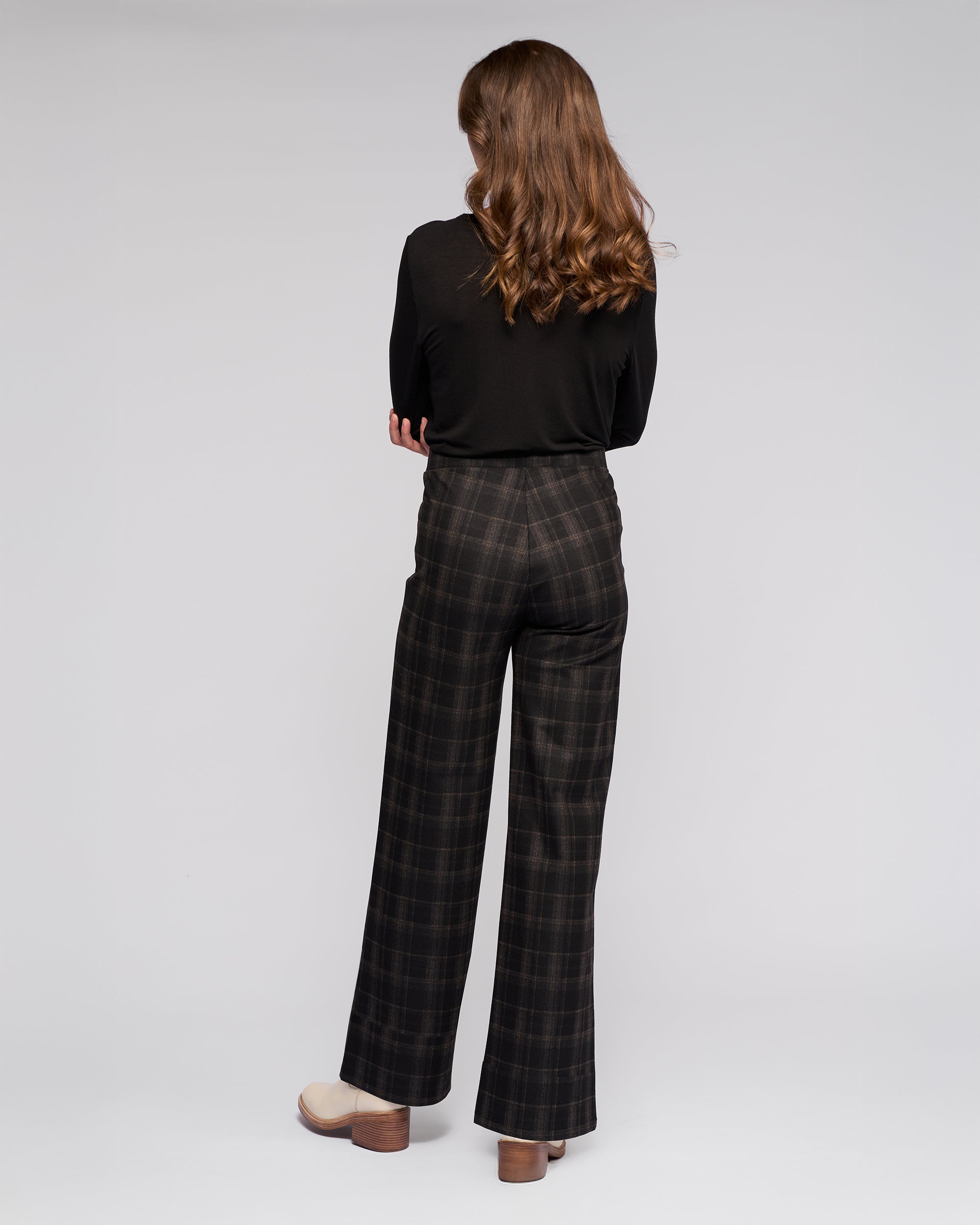 A person with wavy brown hair faces away, wearing Vassalli Loft Pull On - Checkmate high-waisted wide-leg plaid trousers, a black long-sleeve top, and beige platform shoes against a plain light gray background.