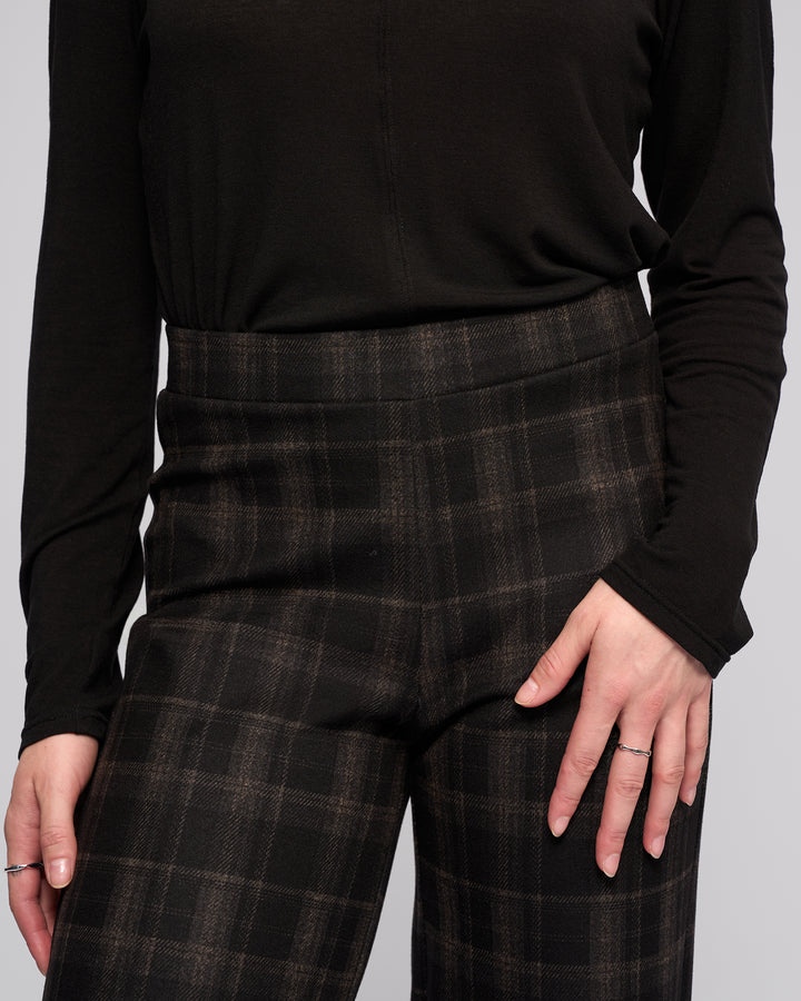 A person wears Vassalli’s Loft Pull On - Checkmate high-waisted, wide-leg pants with a long-sleeve black top, one hand on their hip and the other relaxed, showing simple rings.