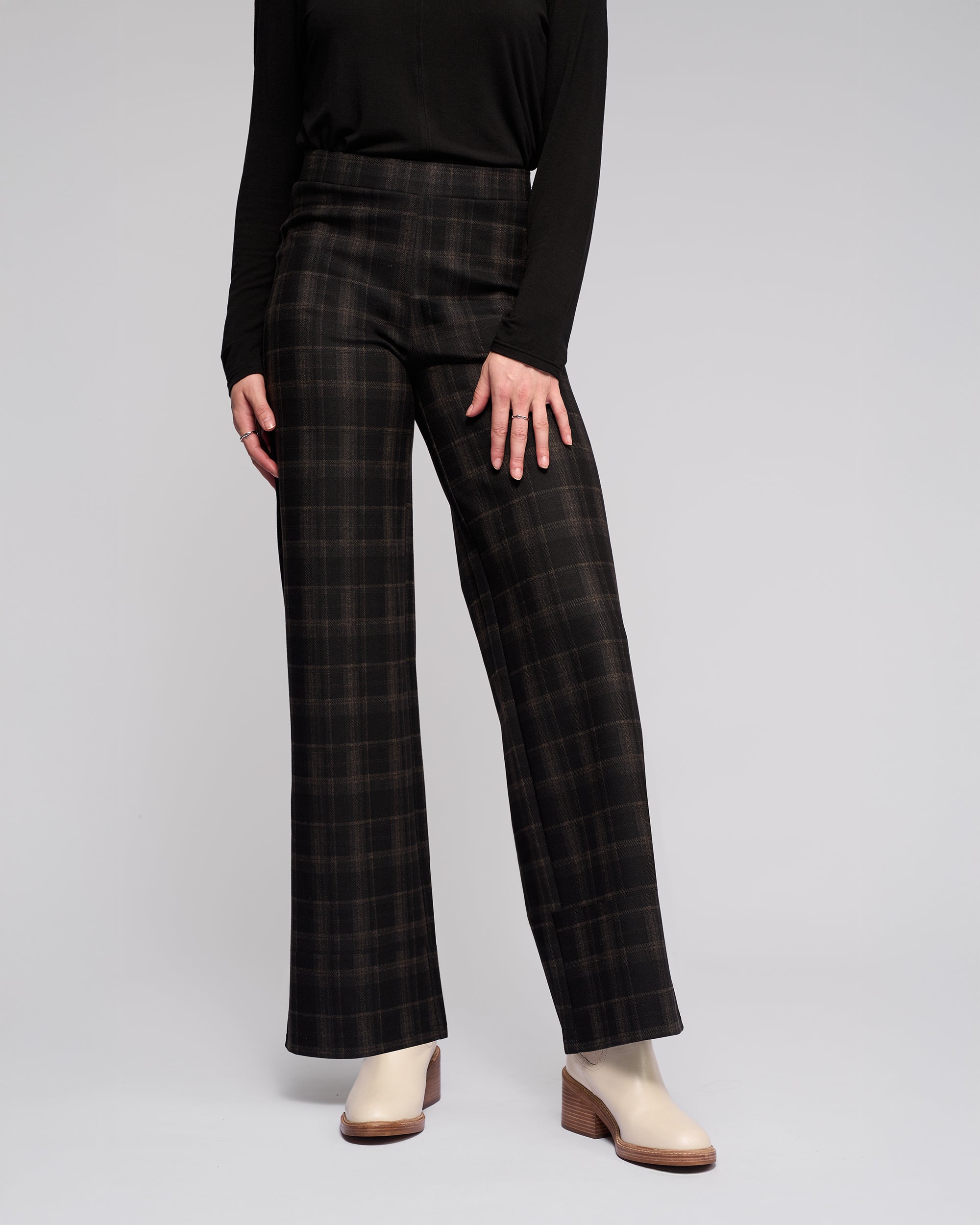 A person models the Vassalli Loft Pull On - Checkmate high-waisted, wide-leg pants with a long-sleeve black top and light-colored heeled ankle boots against a plain light background. The face is not shown.