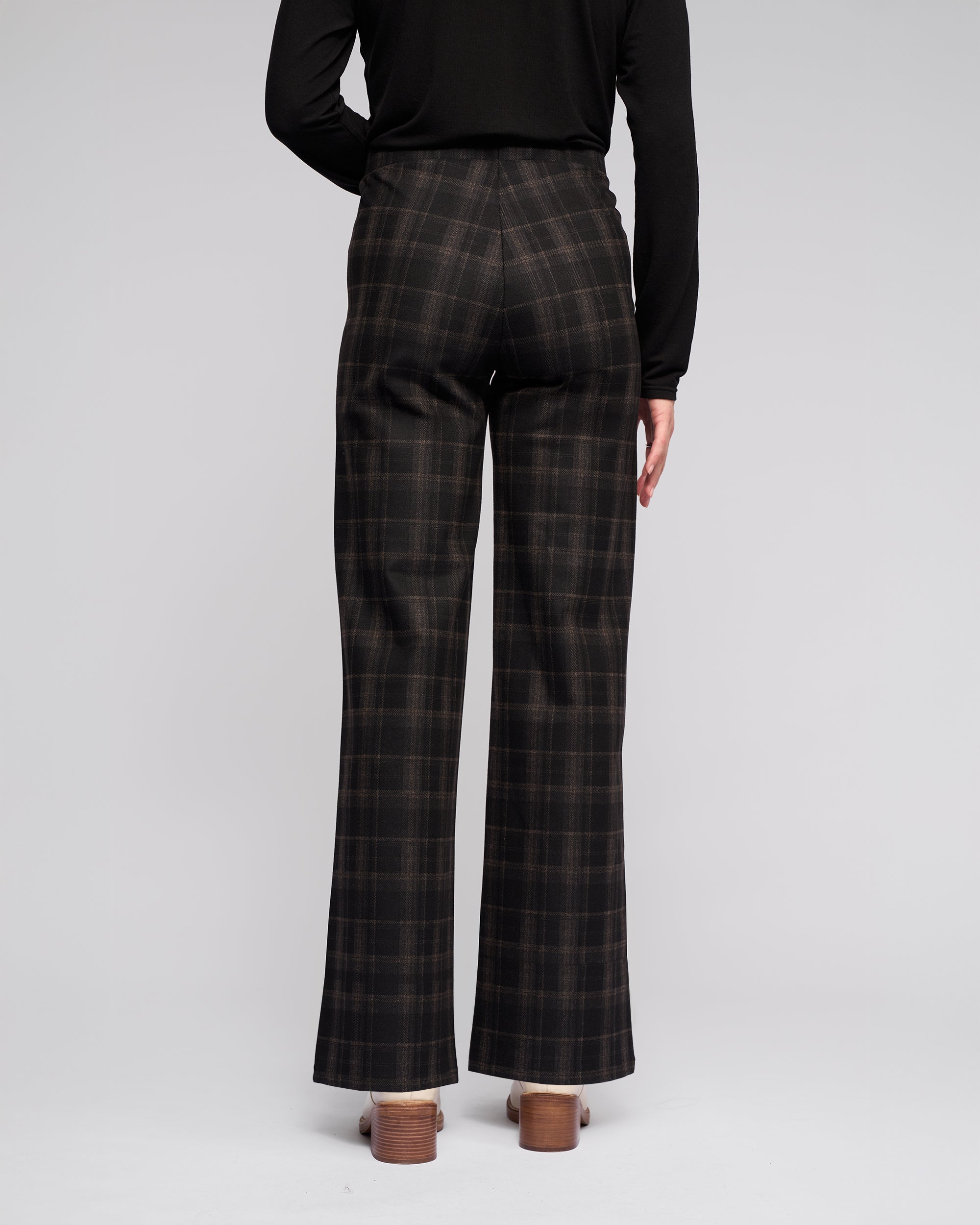 A person stands facing away from the camera, wearing Vassalli Loft Pull On - Checkmate wide-leg pants, a long-sleeve black top, and brown heeled shoes against a plain light background.