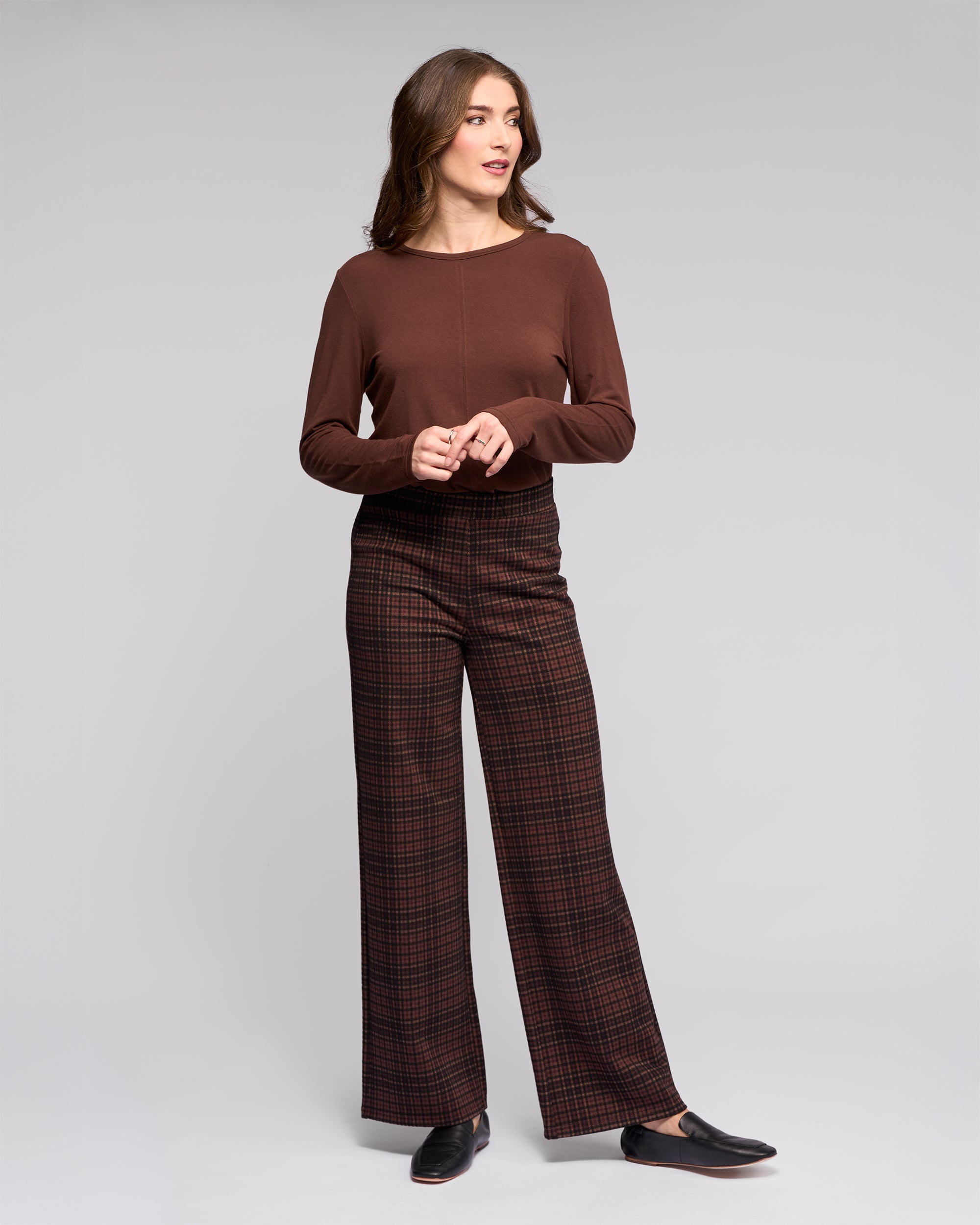 A woman stands against a plain background, wearing the Vassalli Loft Pull On - Cherry stretch knit wide-leg pants and a long-sleeved brown top, with black shoes, looking off to the side with her hands loosely clasped.