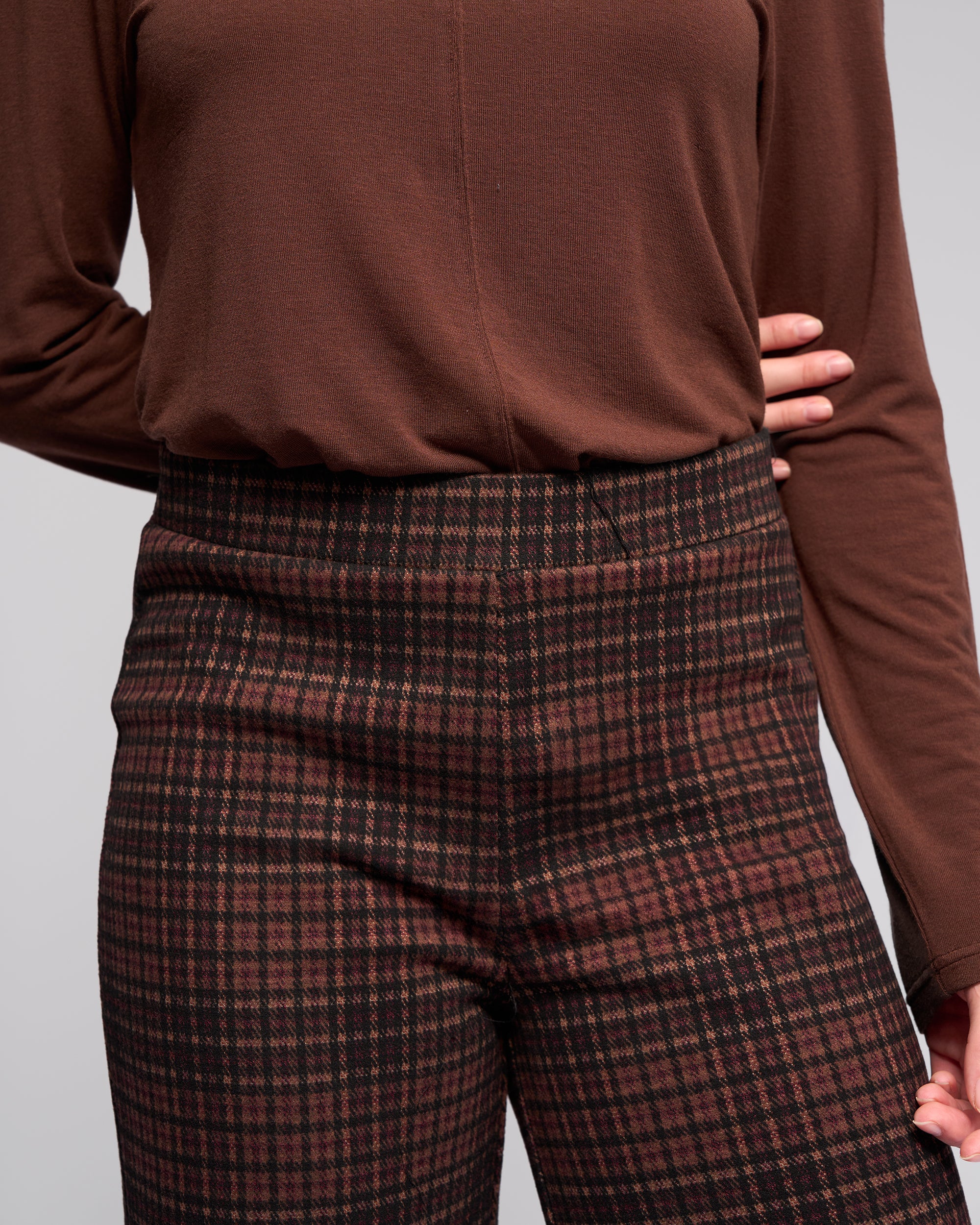 Someone stands against a neutral background with one hand on their hip, wearing the Vassalli Loft Pull On - Cherry pants featuring a high waist, wide-leg fit, plaid pattern, and elasticated waistband.