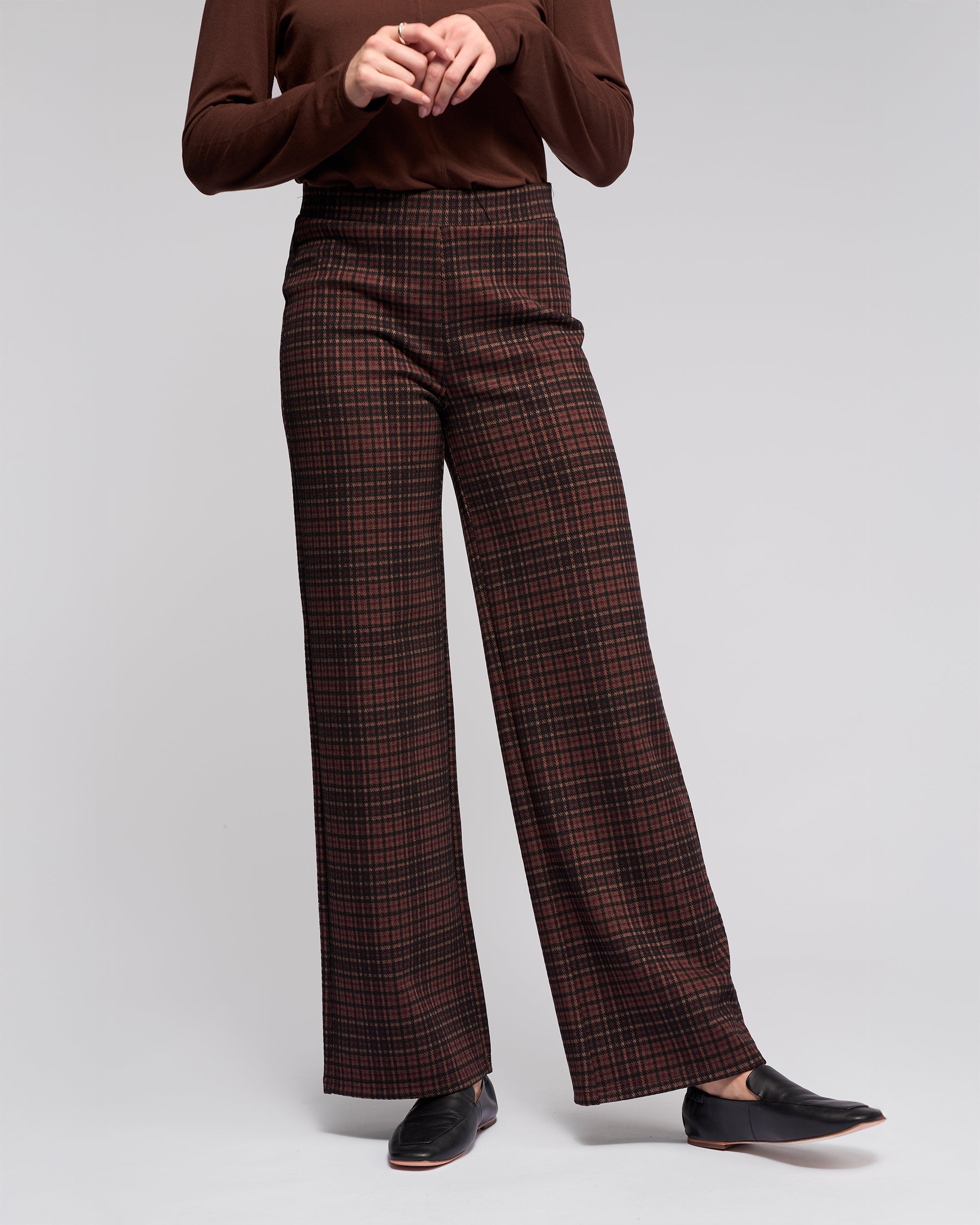 A person wears Vassalli's Loft Pull On - Cherry high-waisted, wide-leg plaid pants with an elastic waistband in dark red and brown, a stretch knit long-sleeve brown top, and black loafers against a plain light background. Their face isn't visible.