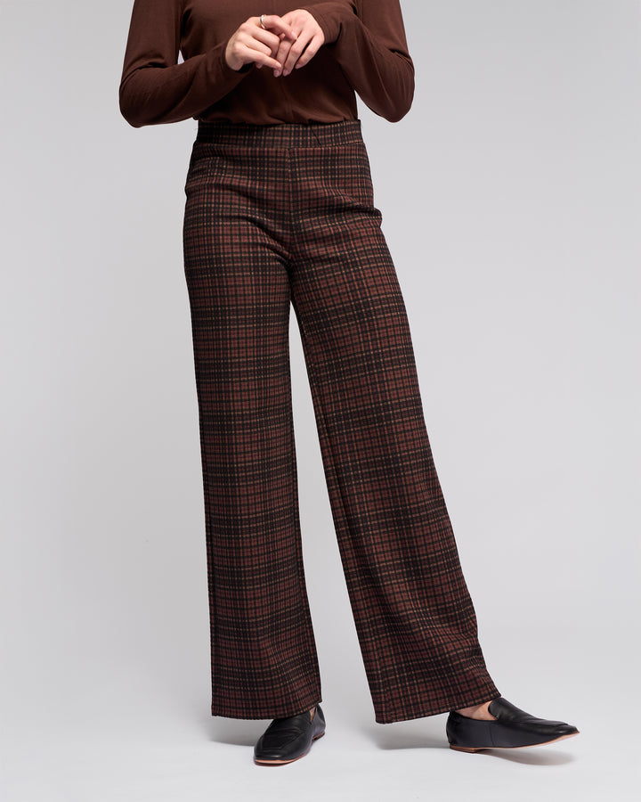 A person wears Vassalli's Loft Pull On - Cherry high-waisted, wide-leg plaid pants with an elastic waistband in dark red and brown, a stretch knit long-sleeve brown top, and black loafers against a plain light background. Their face isn't visible.