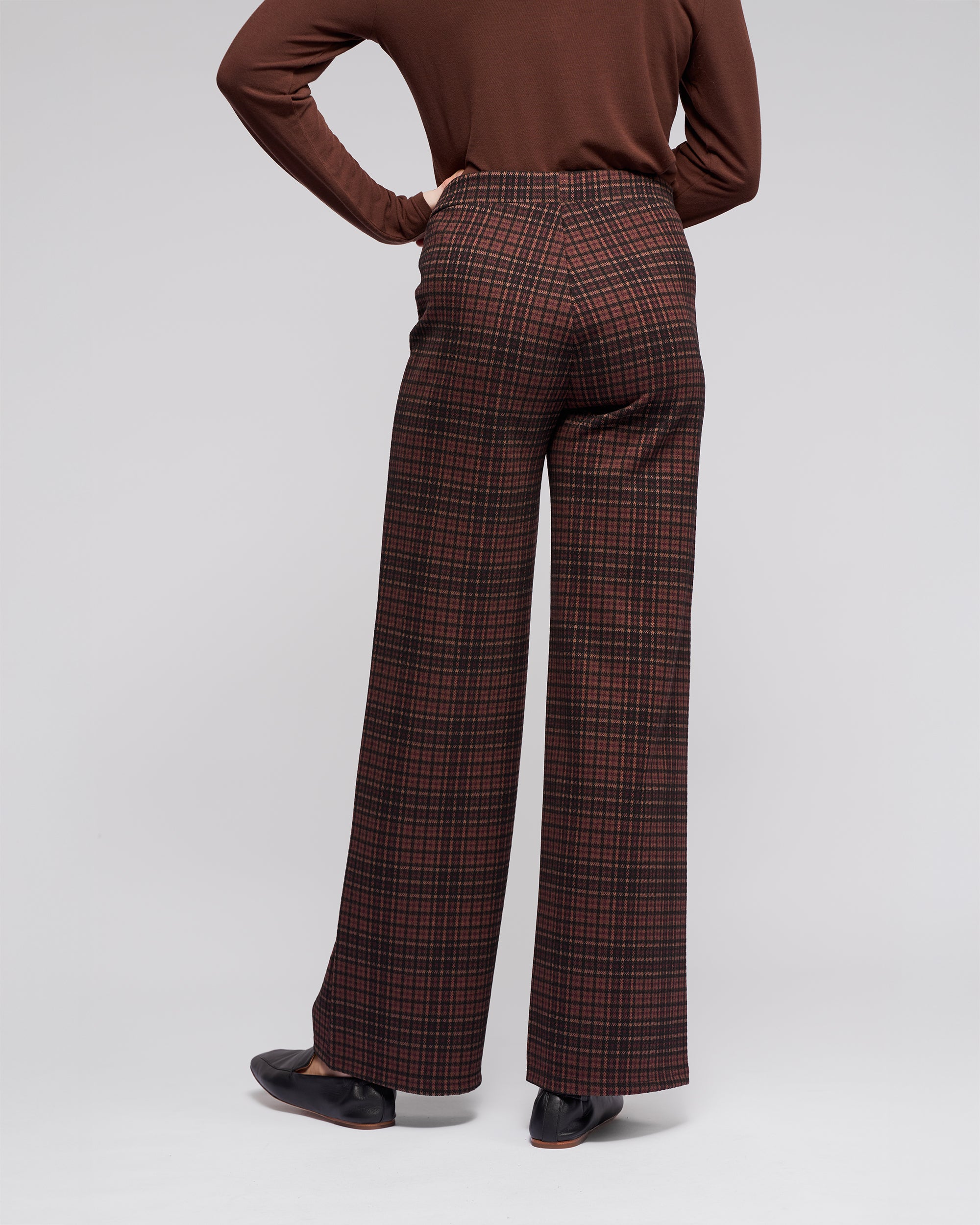A person wears Vassalli's Loft Pull On - Cherry high-waisted, wide-leg plaid trousers in brown and red, styled with a brown long-sleeve top and black shoes, standing with one hand on their hip against a plain background.