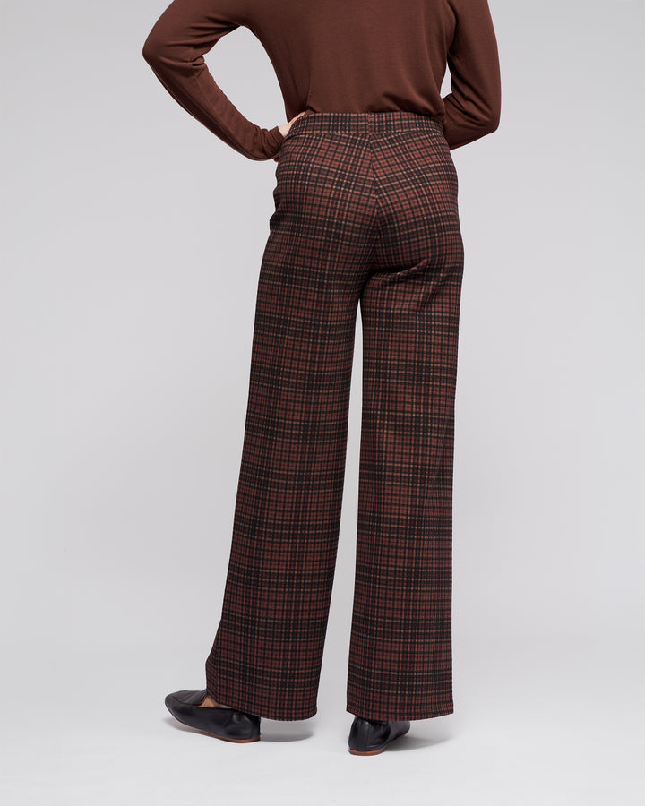 A person wears Vassalli's Loft Pull On - Cherry high-waisted, wide-leg plaid trousers in brown and red, styled with a brown long-sleeve top and black shoes, standing with one hand on their hip against a plain background.