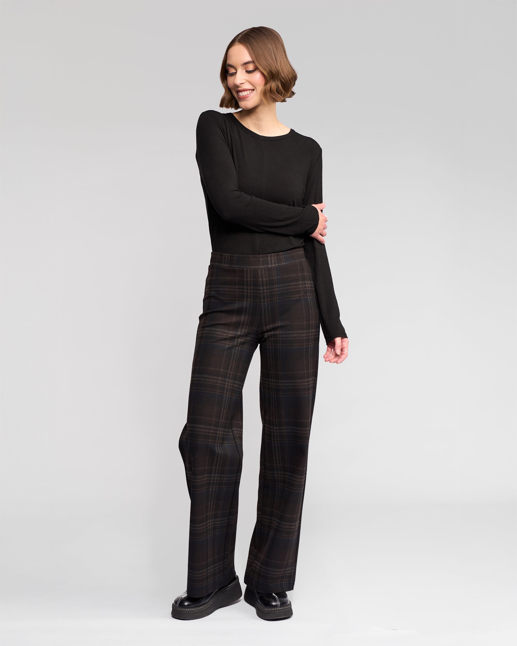 A woman with short brown hair, dressed in a black long-sleeve top and Vassalli Loft Pull On - Hugo wide-leg pants, stands smiling with one arm crossed against a plain light gray background.