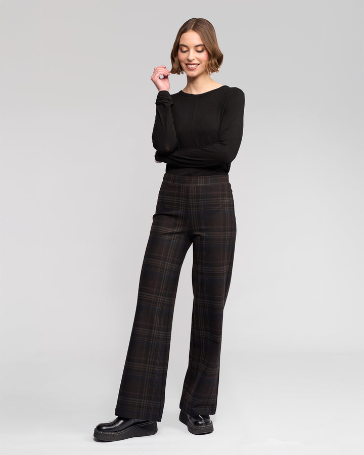 A woman stands against a light gray background, smiling with arms loosely crossed. She wears the Vassalli Loft Pull On - Hugo black long-sleeve top and dark wide-leg plaid stretch ponti pants, paired with black platform shoes.