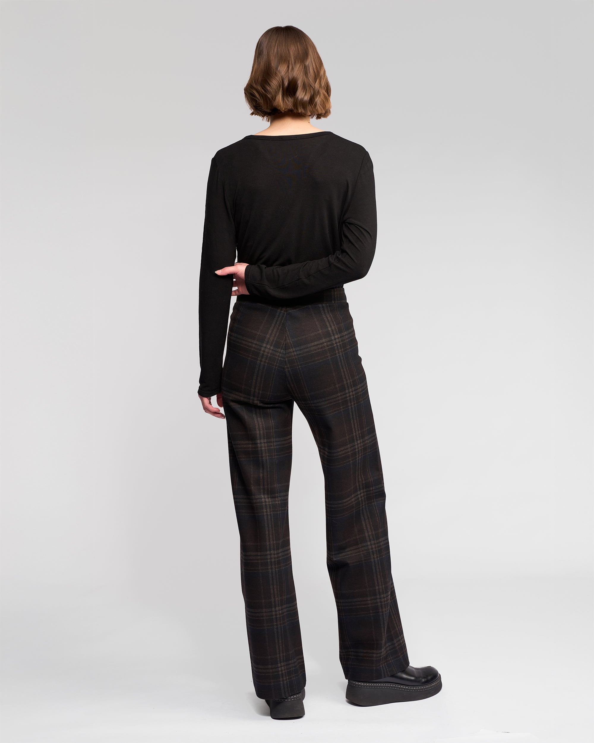 A person with short brown hair, wearing the Vassalli Loft Pull On - Hugo pant and a long-sleeve black top with black shoes, stands facing away from the camera against a plain light gray background.