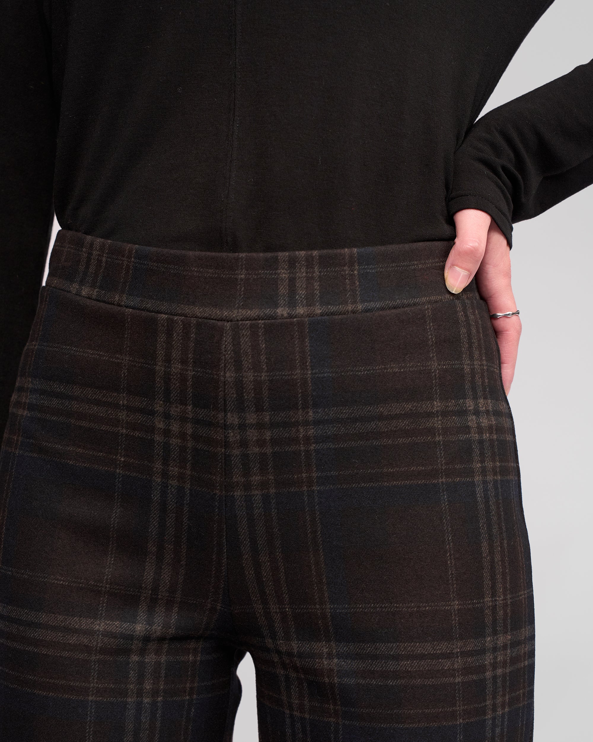 Wearing Vassalli's Loft Pull On - Hugo pants, a person stands with one hand on their hip in high-waisted, dark plaid wide-leg trousers and a black long-sleeve top, showcasing a silver ring.