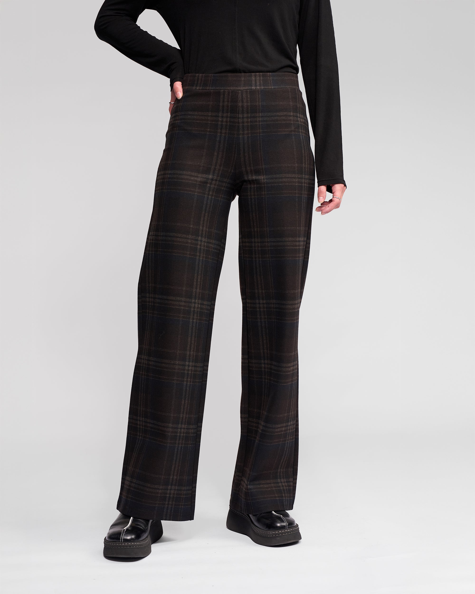 A person wears the Vassalli Loft Pull On - Hugo high-waisted plaid wide-leg pants with a black long-sleeve top and black platform shoes, standing against a plain light gray background.