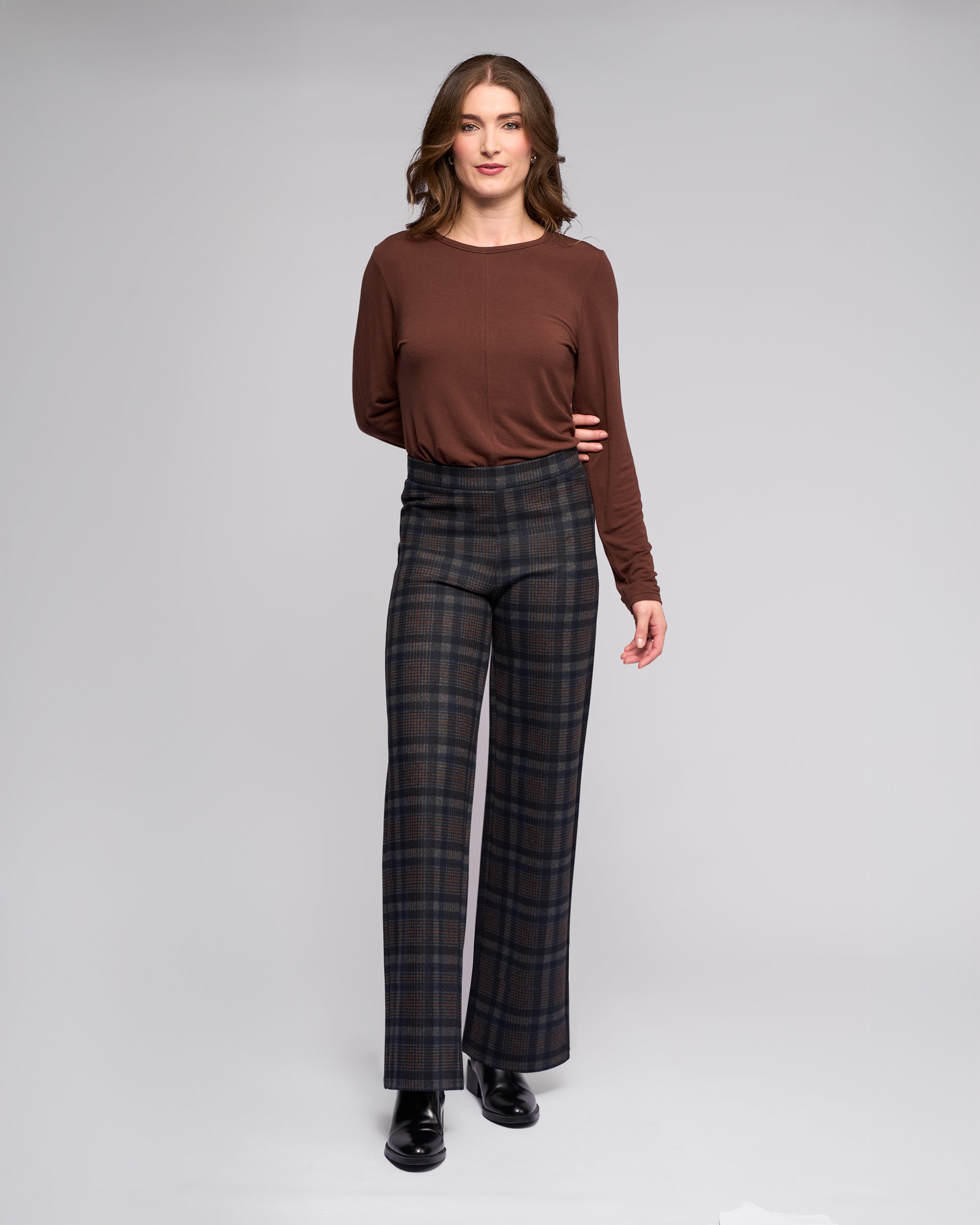 A woman faces forward wearing the Vassalli Loft Pull On - Nomad high-waisted dark plaid wide-leg pants, a long-sleeve brown top, and black ankle boots against a plain light gray background.