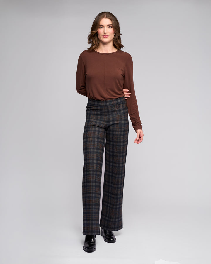 A woman faces forward wearing the Vassalli Loft Pull On - Nomad high-waisted dark plaid wide-leg pants, a long-sleeve brown top, and black ankle boots against a plain light gray background.
