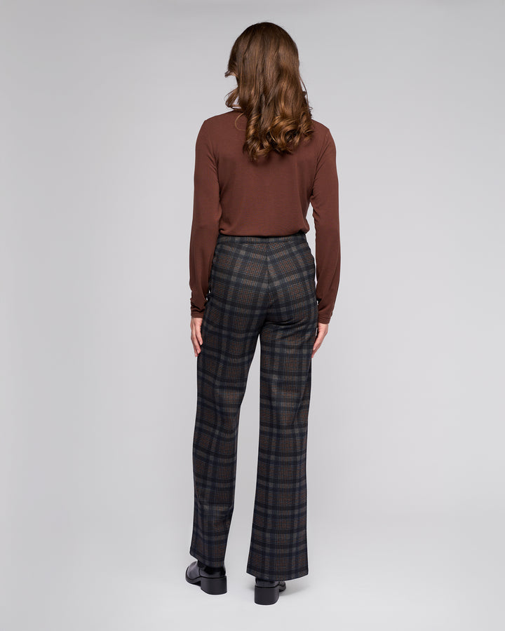 A person with wavy brown hair, in a long-sleeve brown top and Vassalli Loft Pull On - Nomad high-waisted, wide-leg plaid pants, stands facing away against a plain light gray background.
