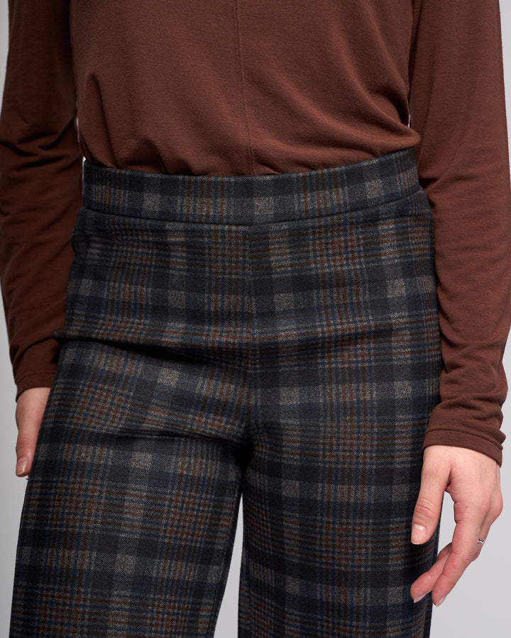 A person is seen from torso to hands wearing the Vassalli Loft Pull On - Nomad high-waisted, wide-leg plaid pants in brown and gray tones, paired with a brown long-sleeve shirt.