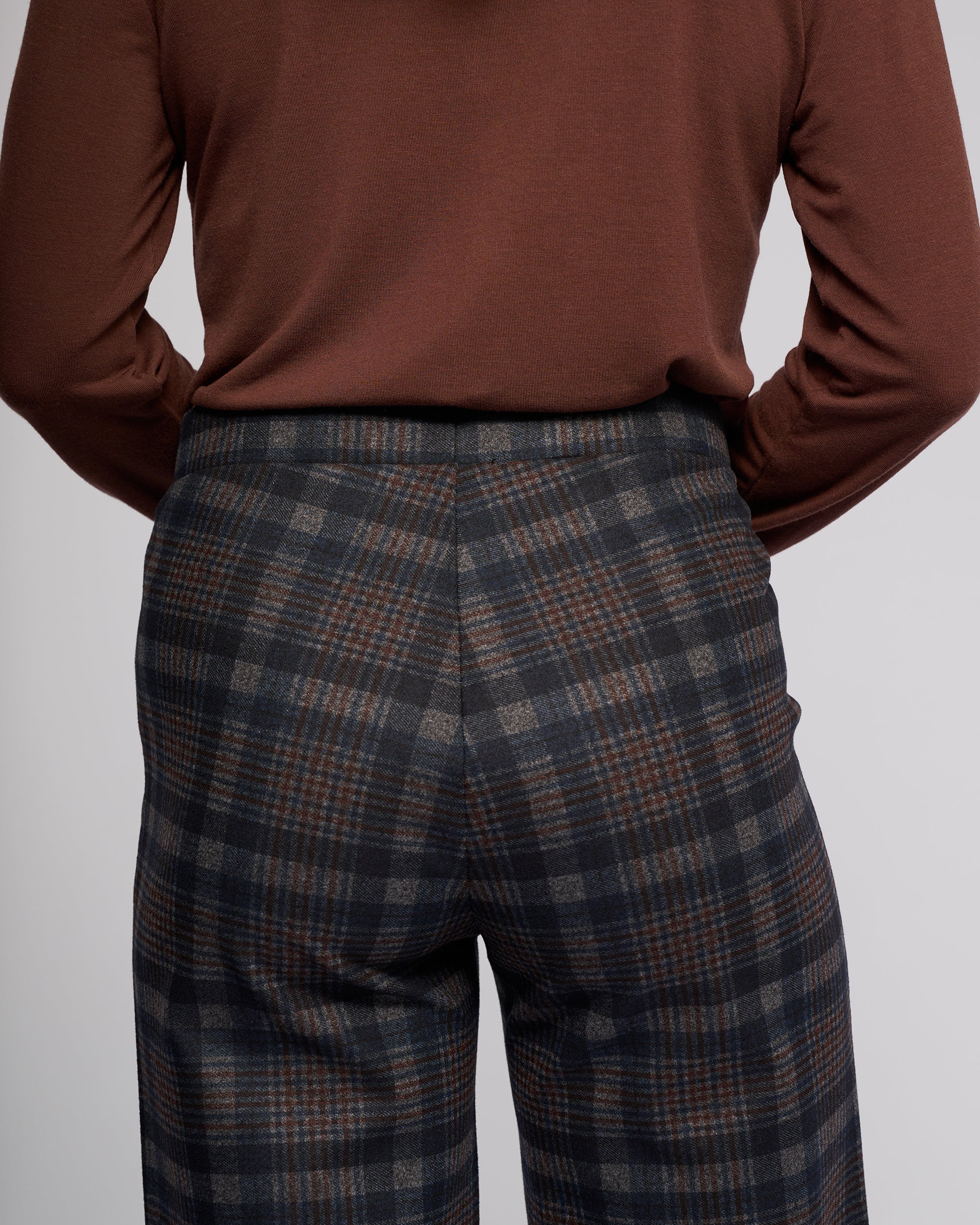 A person is shown from the back wearing Vassalli's Loft Pull On - Nomad: high-waisted, wide-leg plaid trousers in gray, black, and brown with a comfy pull-on design, paired with a brown long-sleeve top.