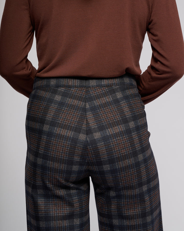 A person is shown from the back wearing Vassalli's Loft Pull On - Nomad: high-waisted, wide-leg plaid trousers in gray, black, and brown with a comfy pull-on design, paired with a brown long-sleeve top.