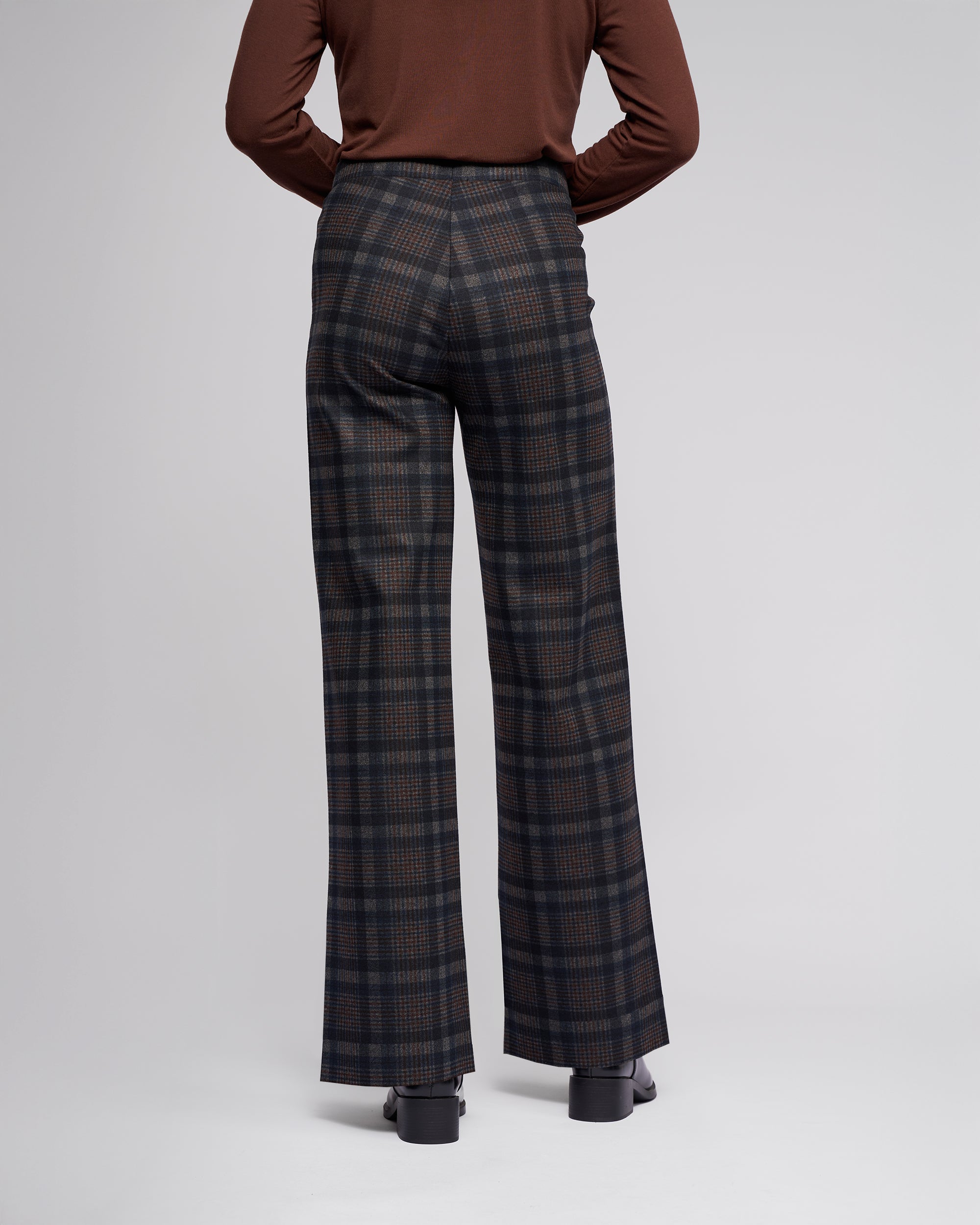 A person wearing the Vassalli Loft Pull On - Nomad, a brown long-sleeve top and high-waisted wide-leg pant in stretch ponti fabric, is seen from the back against a plain light grey background.