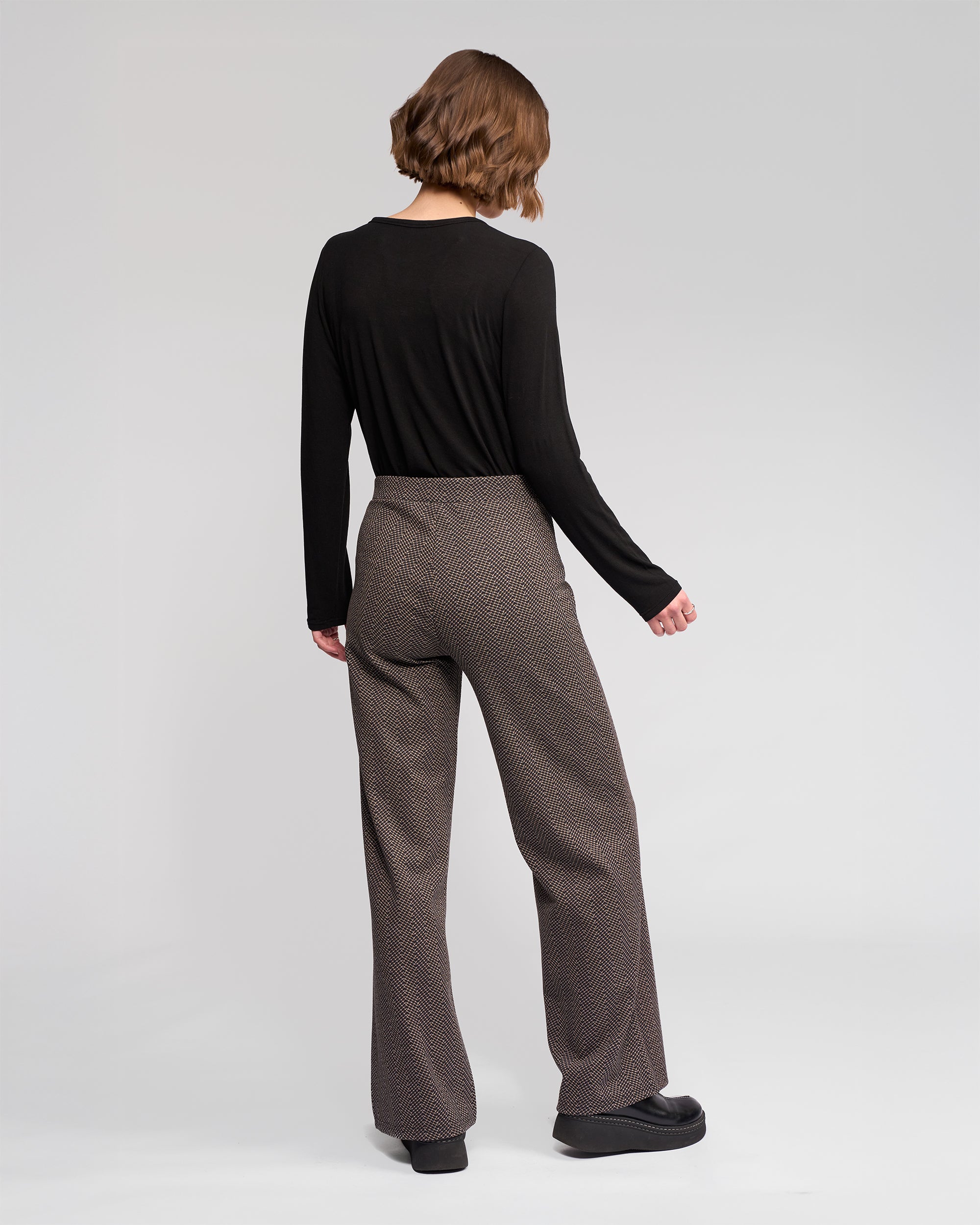 A person with short light brown hair, in a long-sleeved black top and Vassalli Loft Pull On - Arc high-waisted, wide-leg patterned brown trousers with an elasticated waistband, stands facing away against a plain light background.