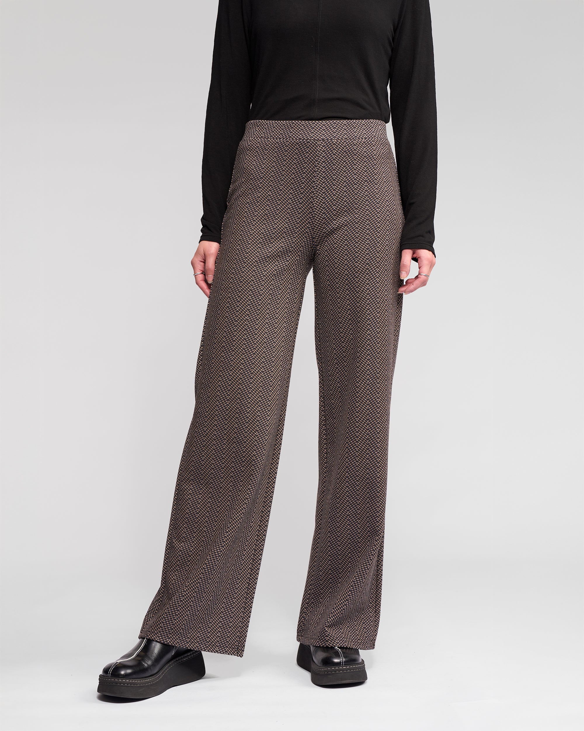 A person wearing the Vassalli Loft Pull On - Arc brown wide-leg pant with an elasticated waistband, a black long-sleeve top, and black platform shoes stands against a plain light gray background.