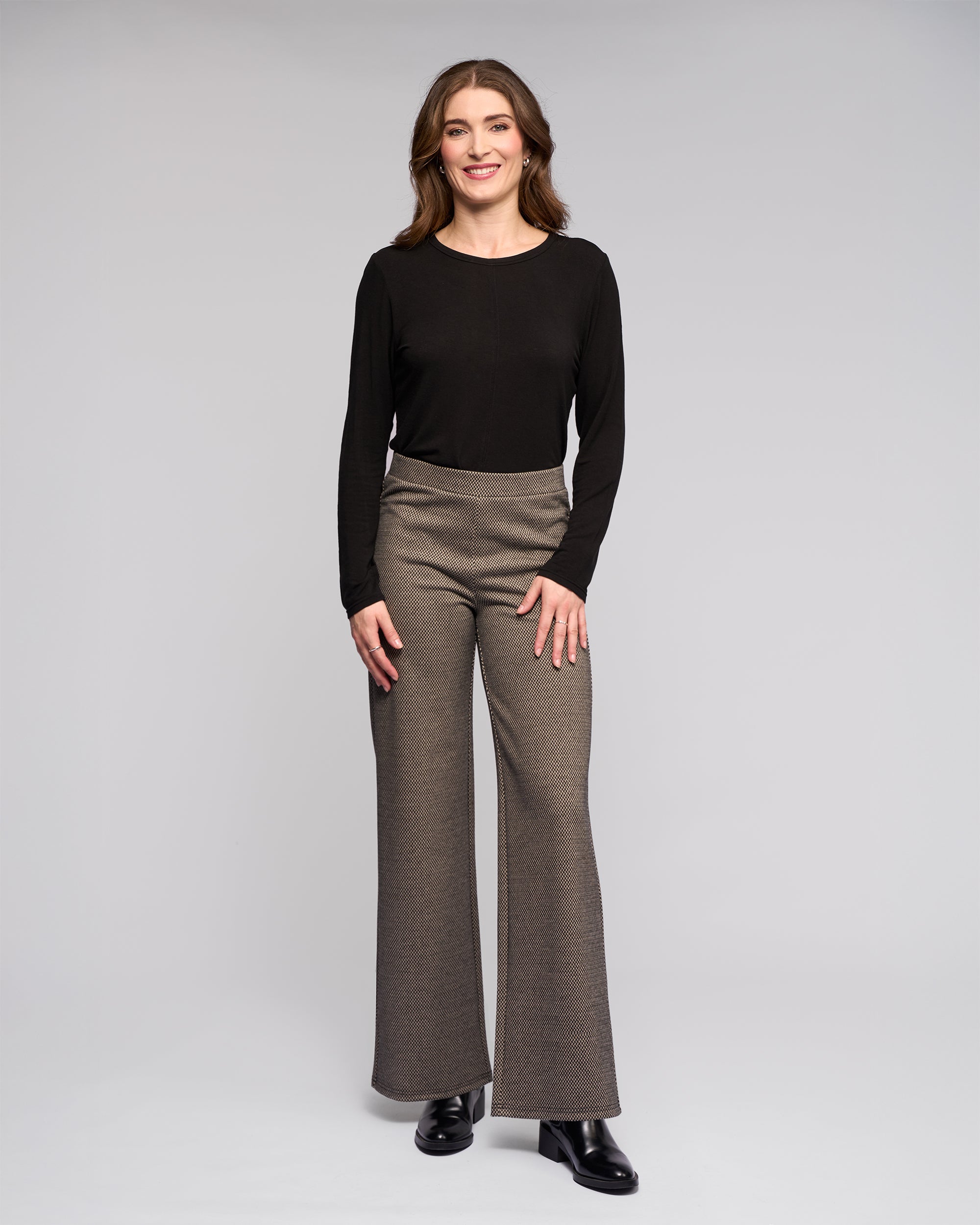 A woman with wavy brown hair, dressed in the Vassalli Loft Pull On - Knox brown high-waisted wide-leg pants and a black long-sleeve top, smiles while standing against a plain light gray background.