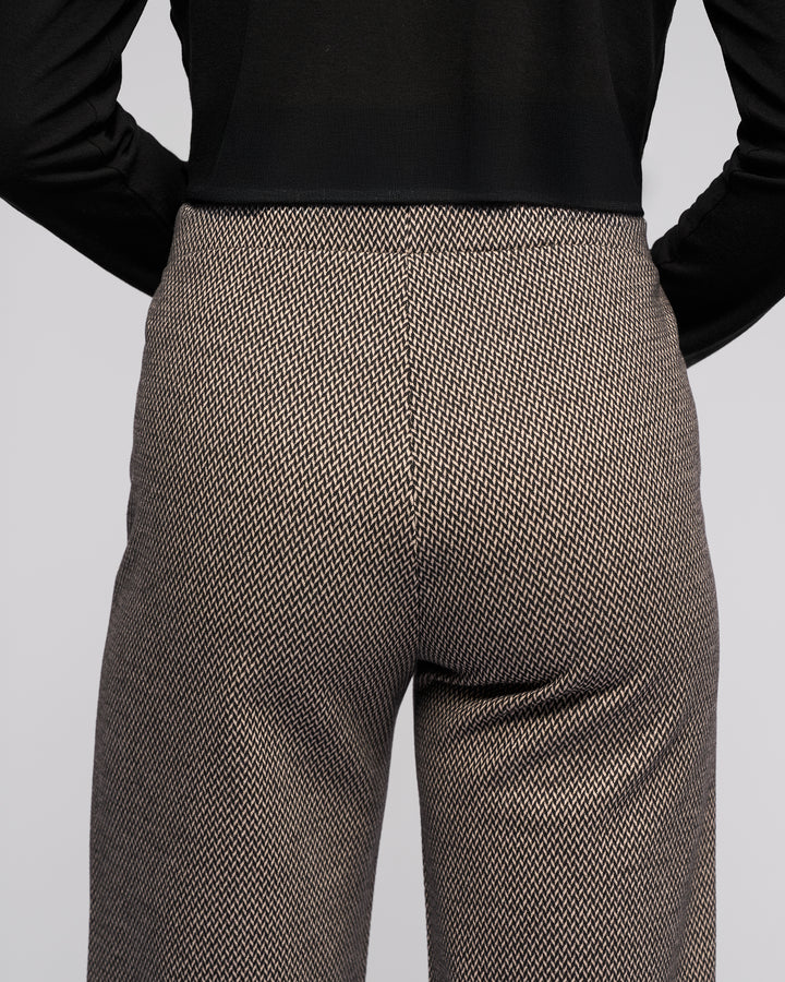 A person wearing the Vassalli Loft Pull On - Knox, a high-waisted, wide-leg brown and white houndstooth pant, paired with a black long-sleeve top, shown from the back with hands on hips.